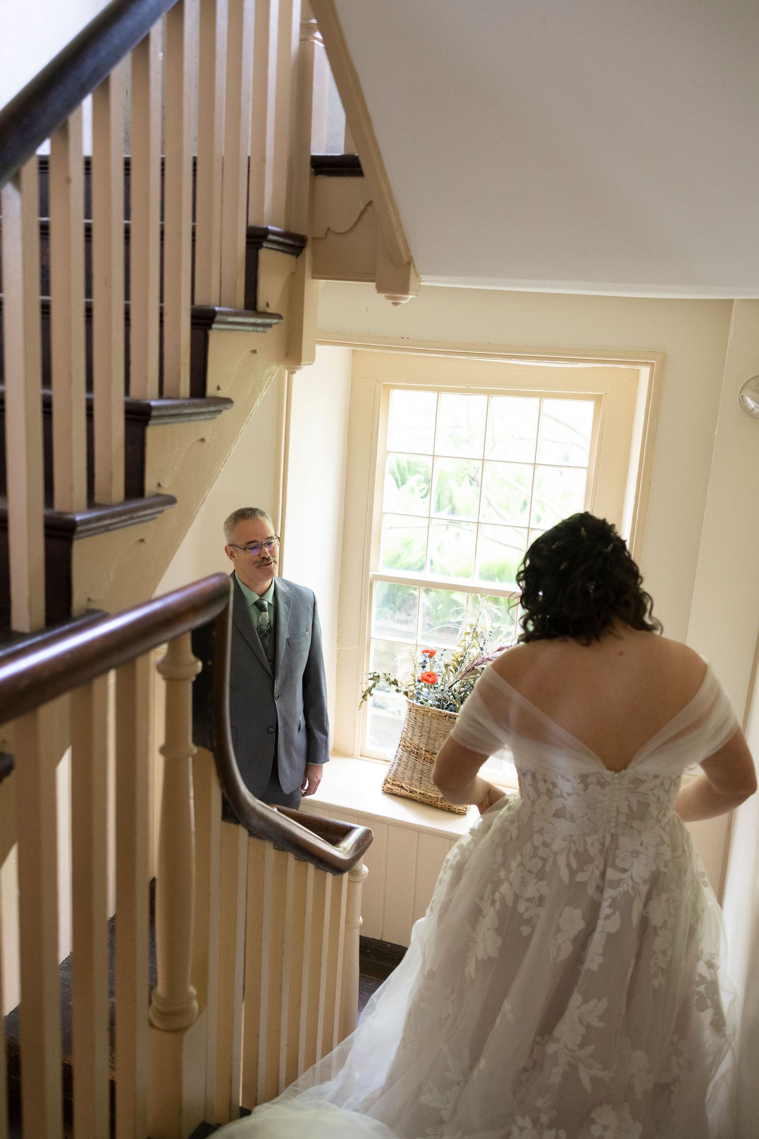 Bride in lace off-shoulder gown descends staircase as groom in gray suit watches during wedding day first look.