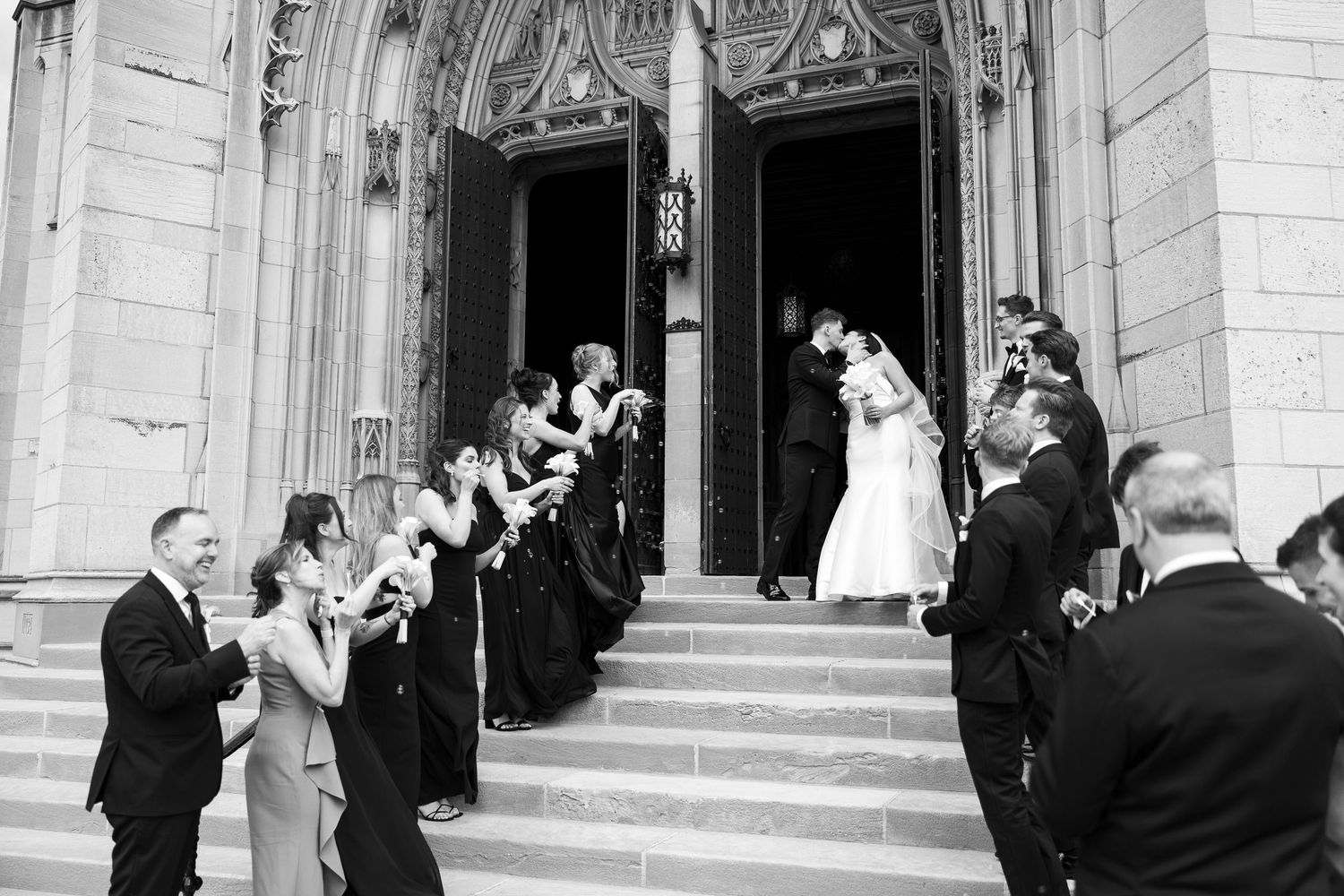 Black and white photo of a bride and groom exiting a grand church, greeted by cheering wedding party on the steps.