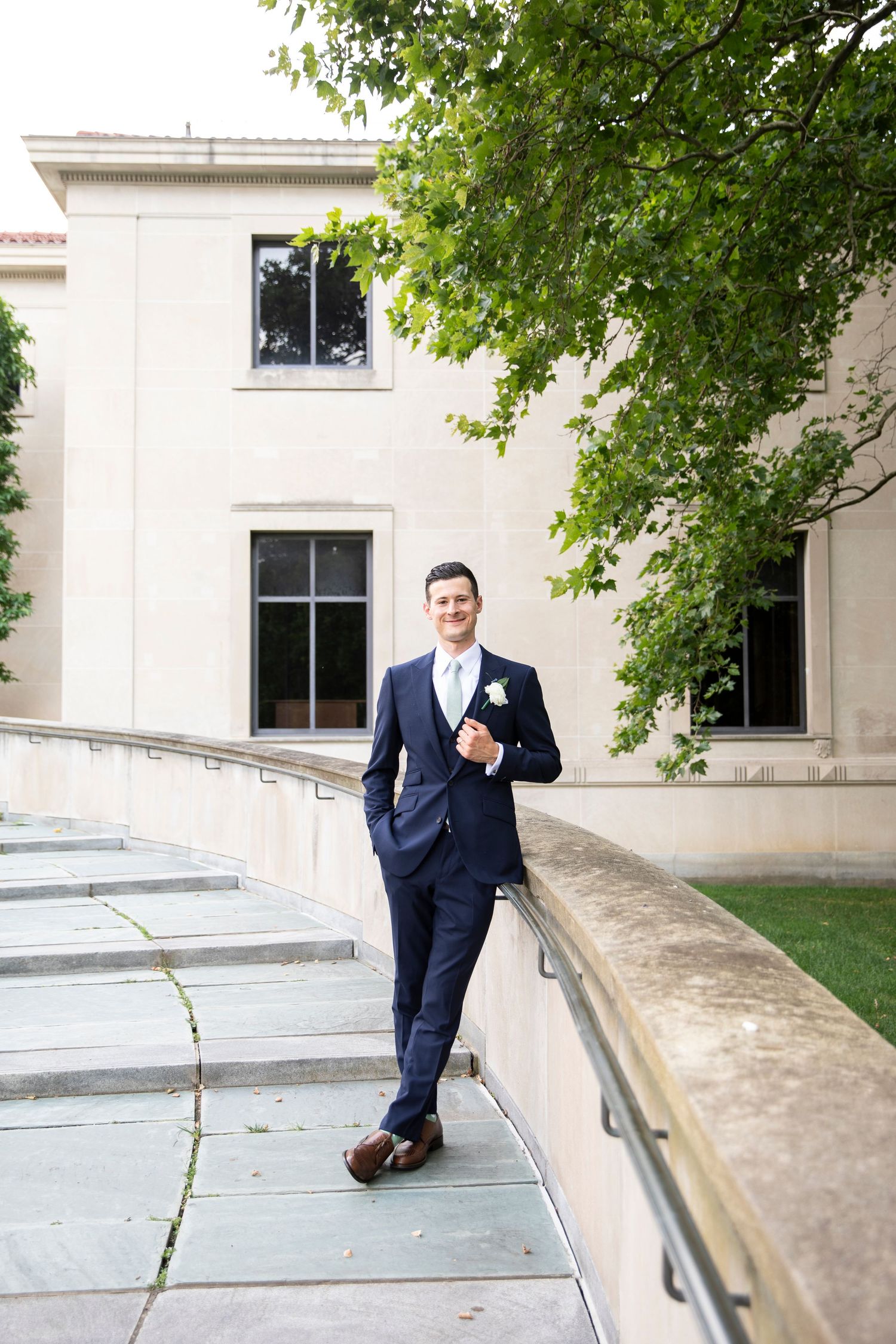 Groom in navy suit poses casually against stone railing of classic building steps.