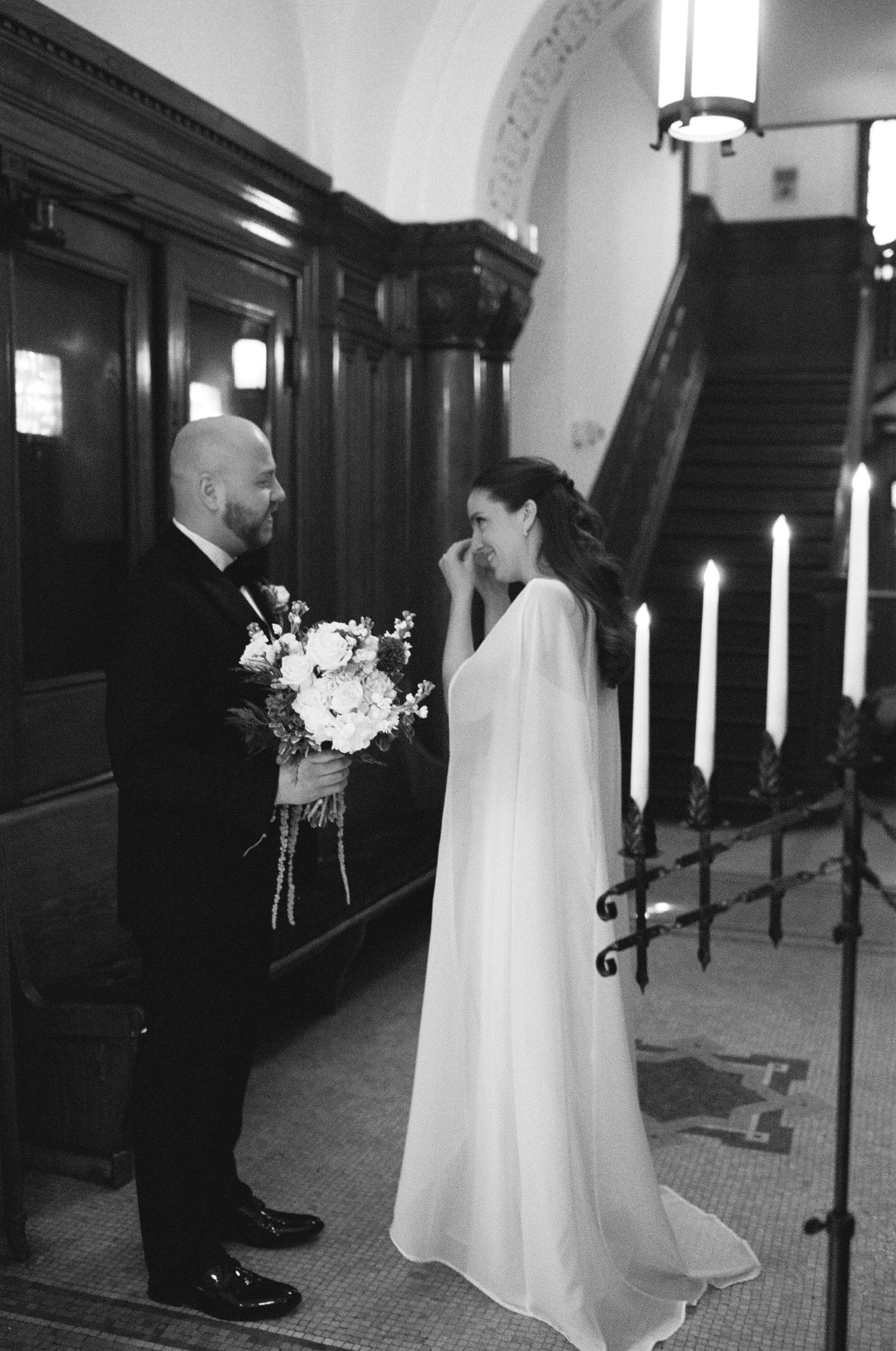 Black and white photo of emotional bride wiping tears as groom holds bouquet during intimate wedding ceremony indoors.