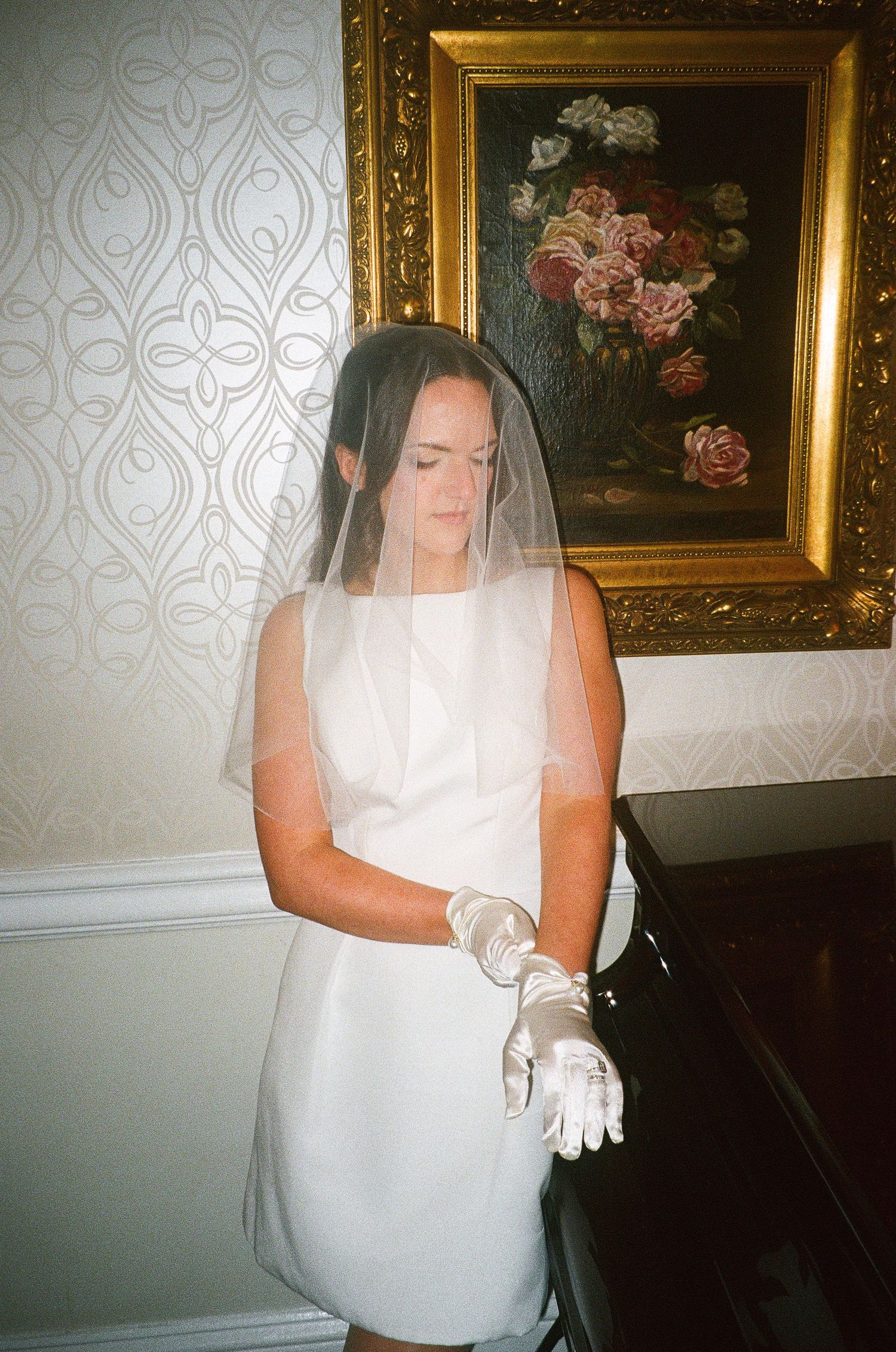 A bride in a sleeveless white dress adjusts her gloves near decorative wall art.