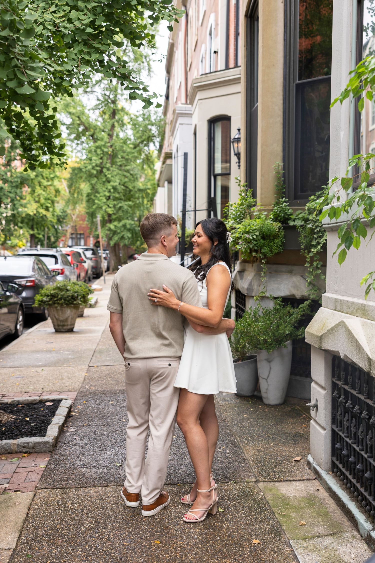 Romantic couple embracing on a tree-lined sidewalk outside historic brownstone buildings.