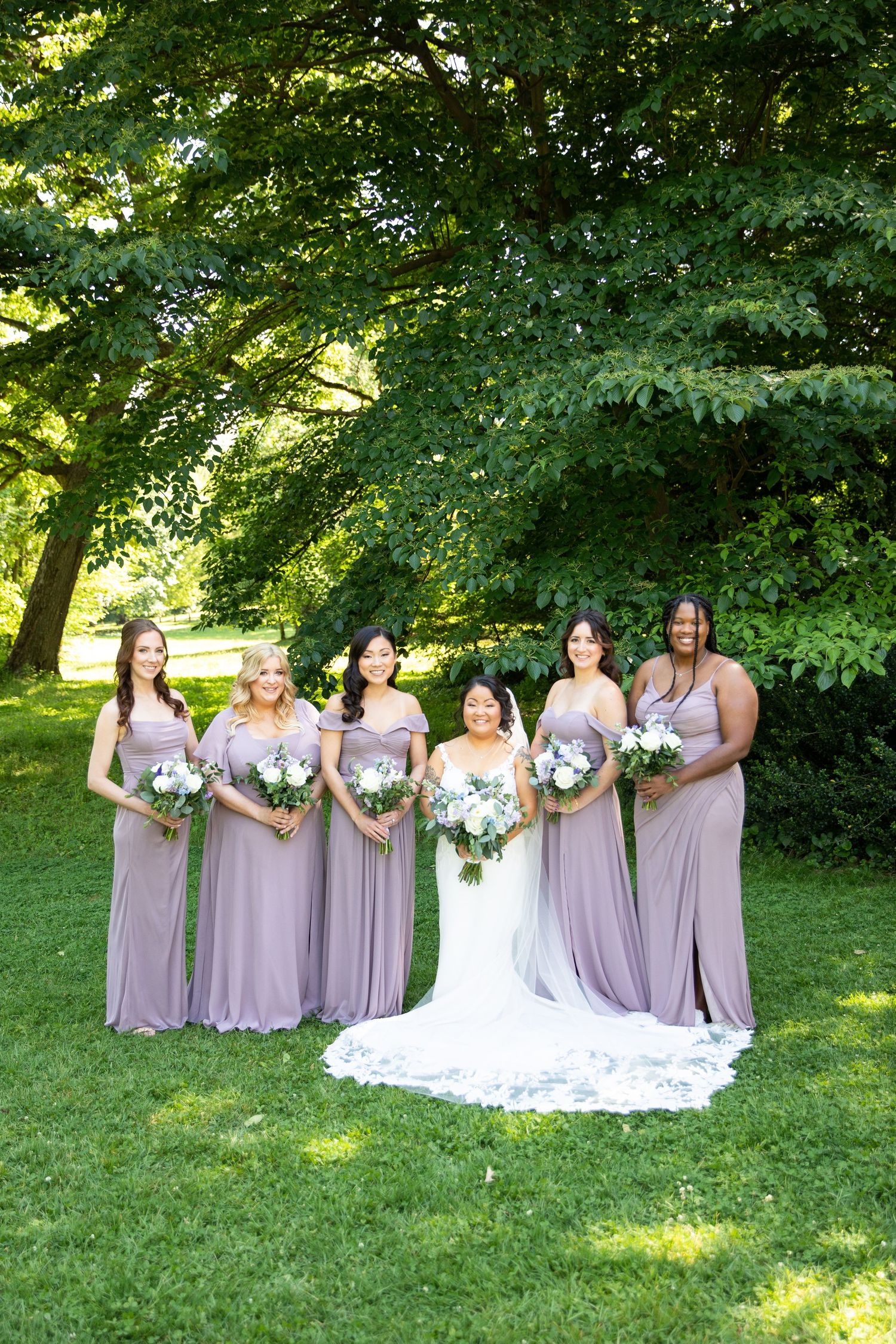 Bridesmaids in flowing lavender dresses pose with bride in white gown on sunny lawn.
