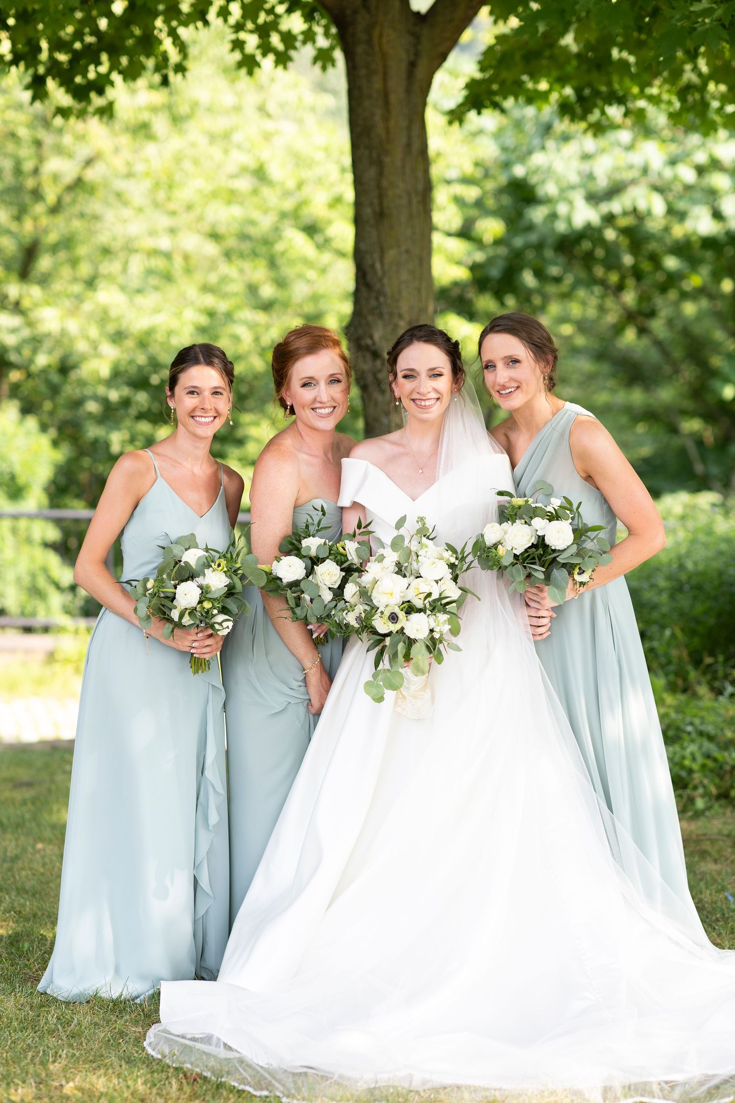 Bridesmaids in pale blue dresses gather around bride holding white bouquets outdoors.