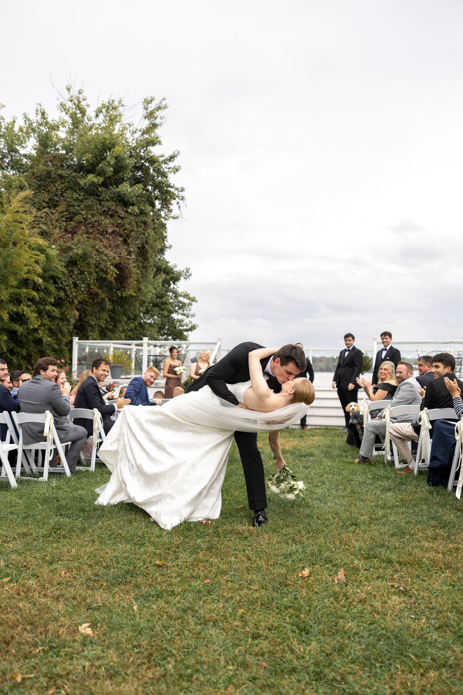 Wedding couple performs dramatic dip pose while guests watch from white chairs on grassy lawn.