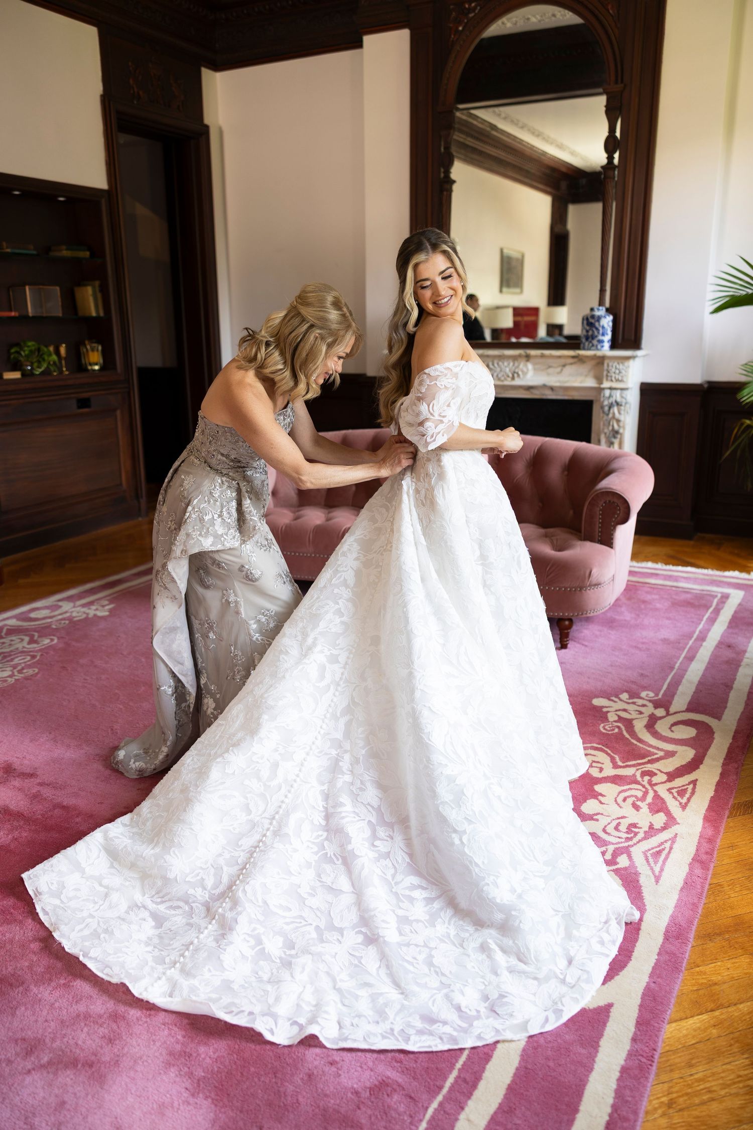 Two women in wedding dresses twirl together on a pink carpet in an elegant room.