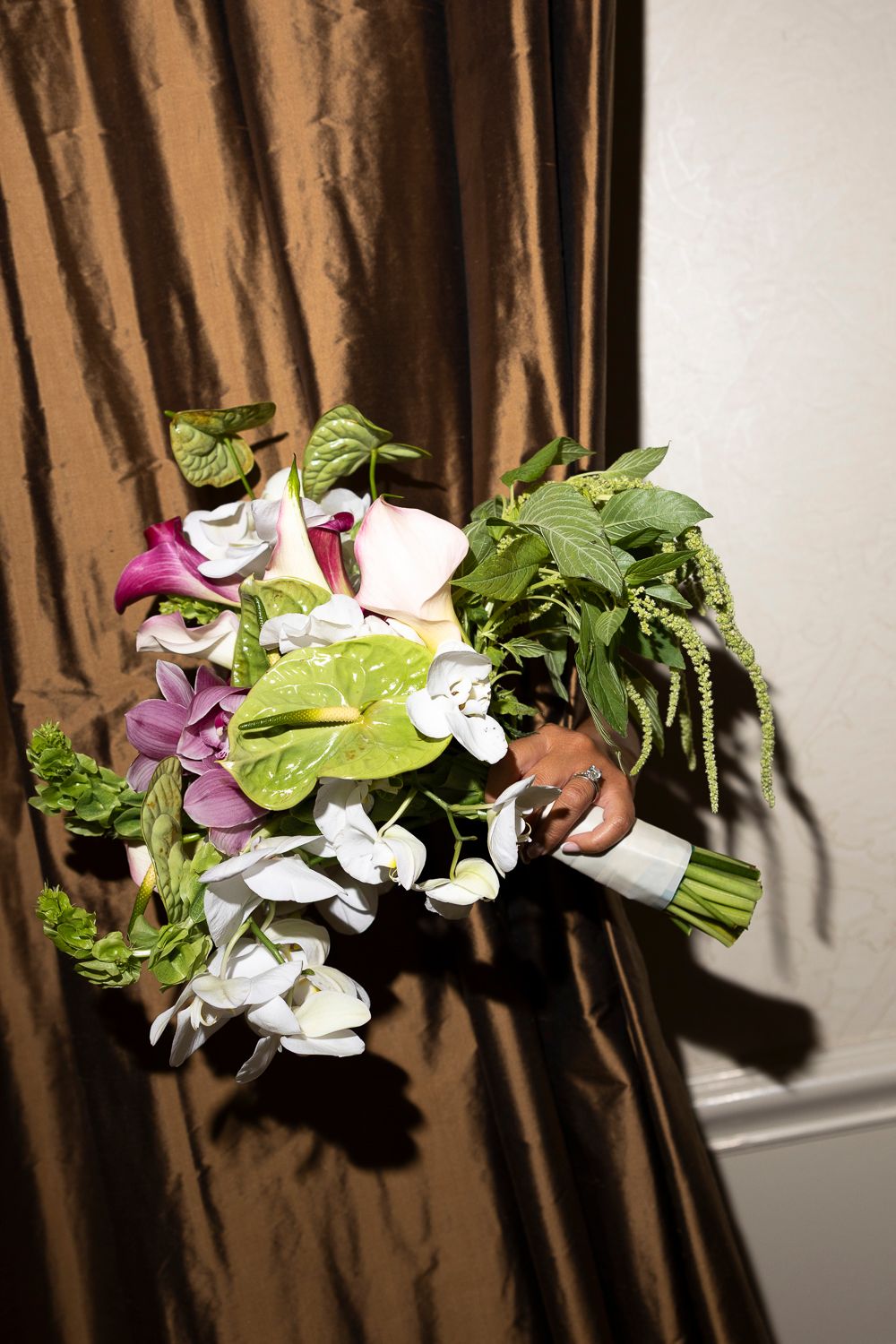 A bridal bouquet with white, pink, and purple flowers, green anthurium leaves, and trailing greenery against a brown satin curtain.