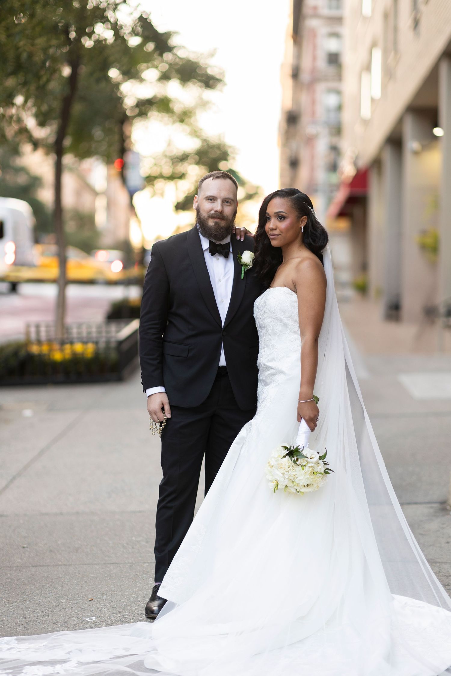 Elegant wedding portrait of couple on city sidewalk with white floral bouquet at sunset.