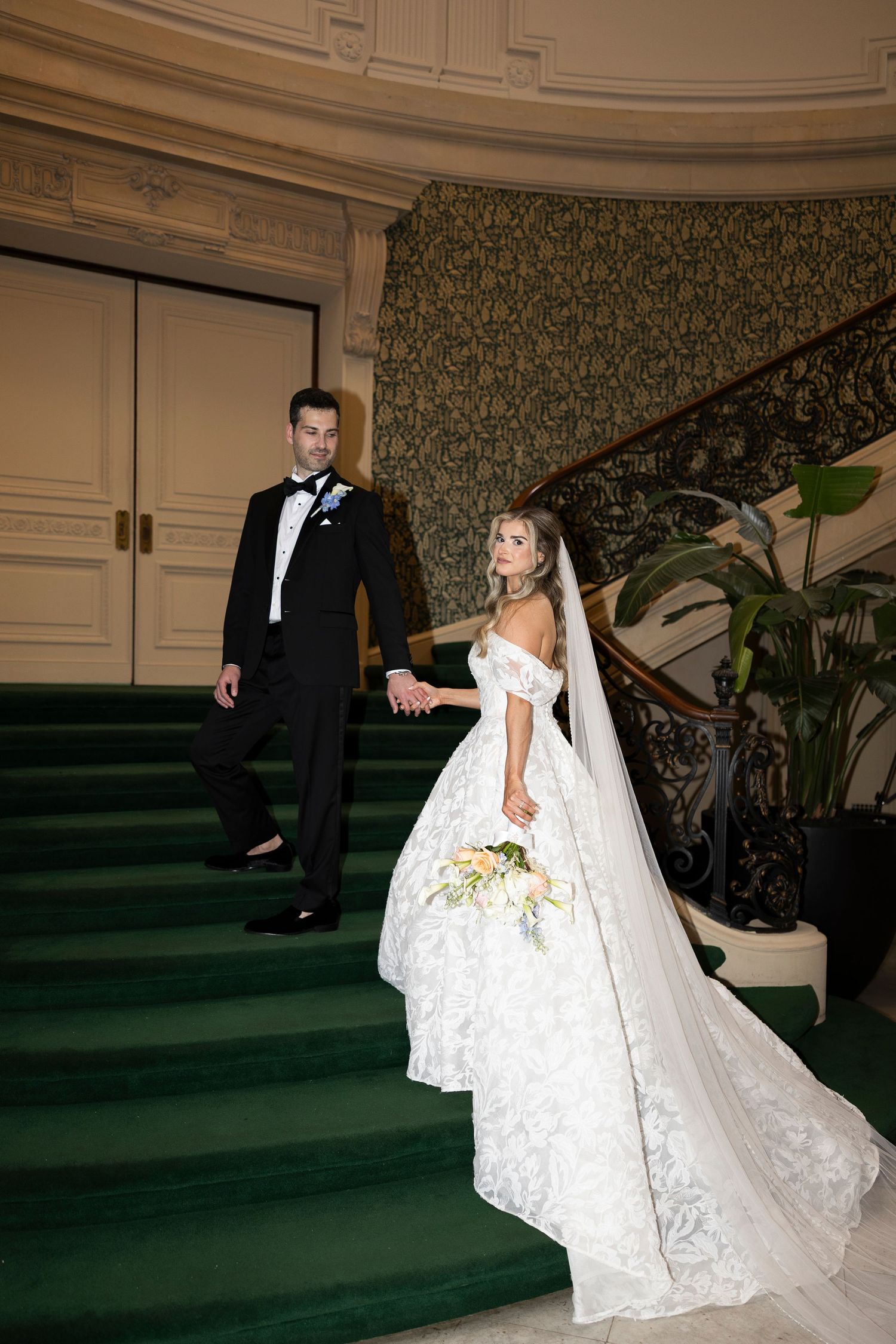 Bride in flowing lace gown poses on elegant green carpeted staircase.