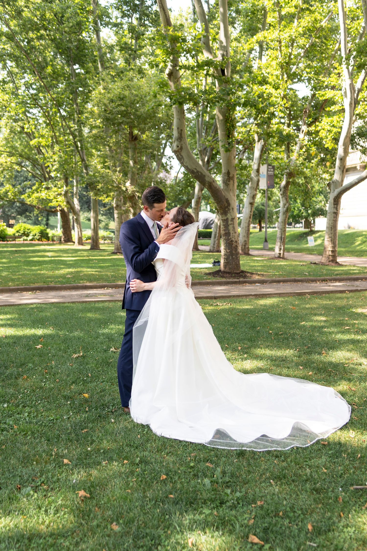 Wedding couple shares intimate moment on sunlit lawn surrounded by mature trees.