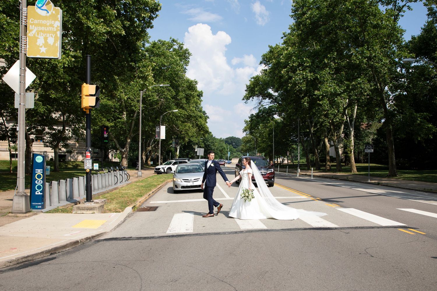 Wedding couple walks along tree-lined street with flowing white dress trailing behind.