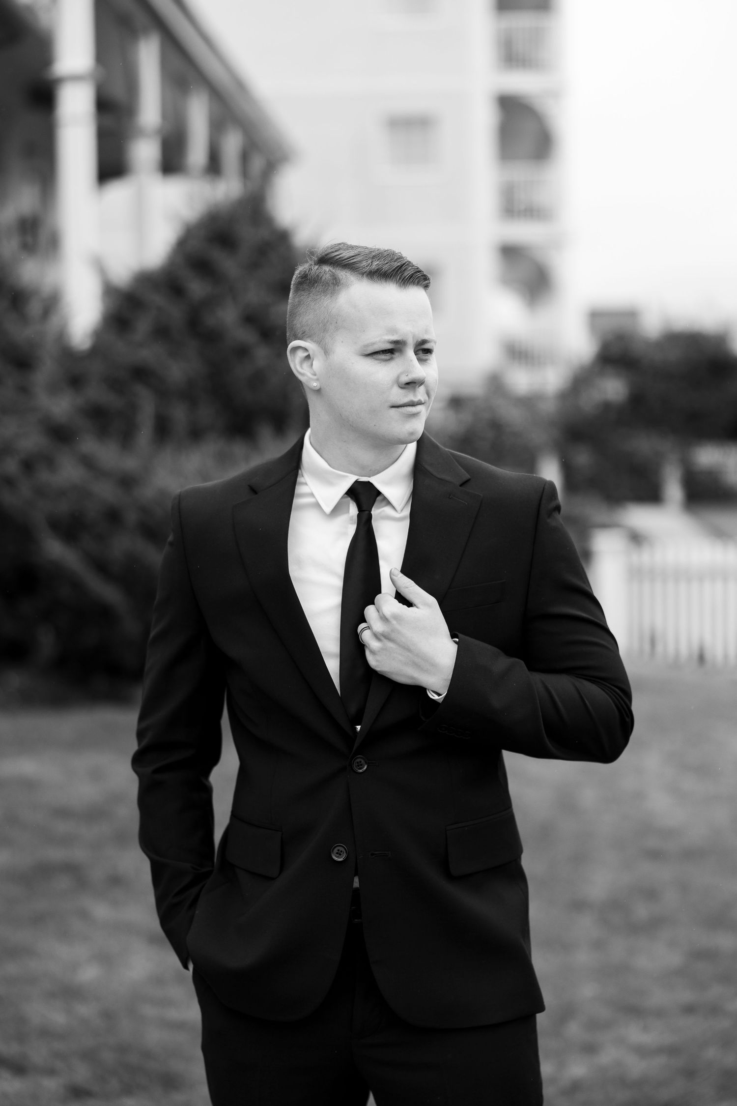 A groom in a black suit adjusts his tie during an outdoor wedding photoshoot.