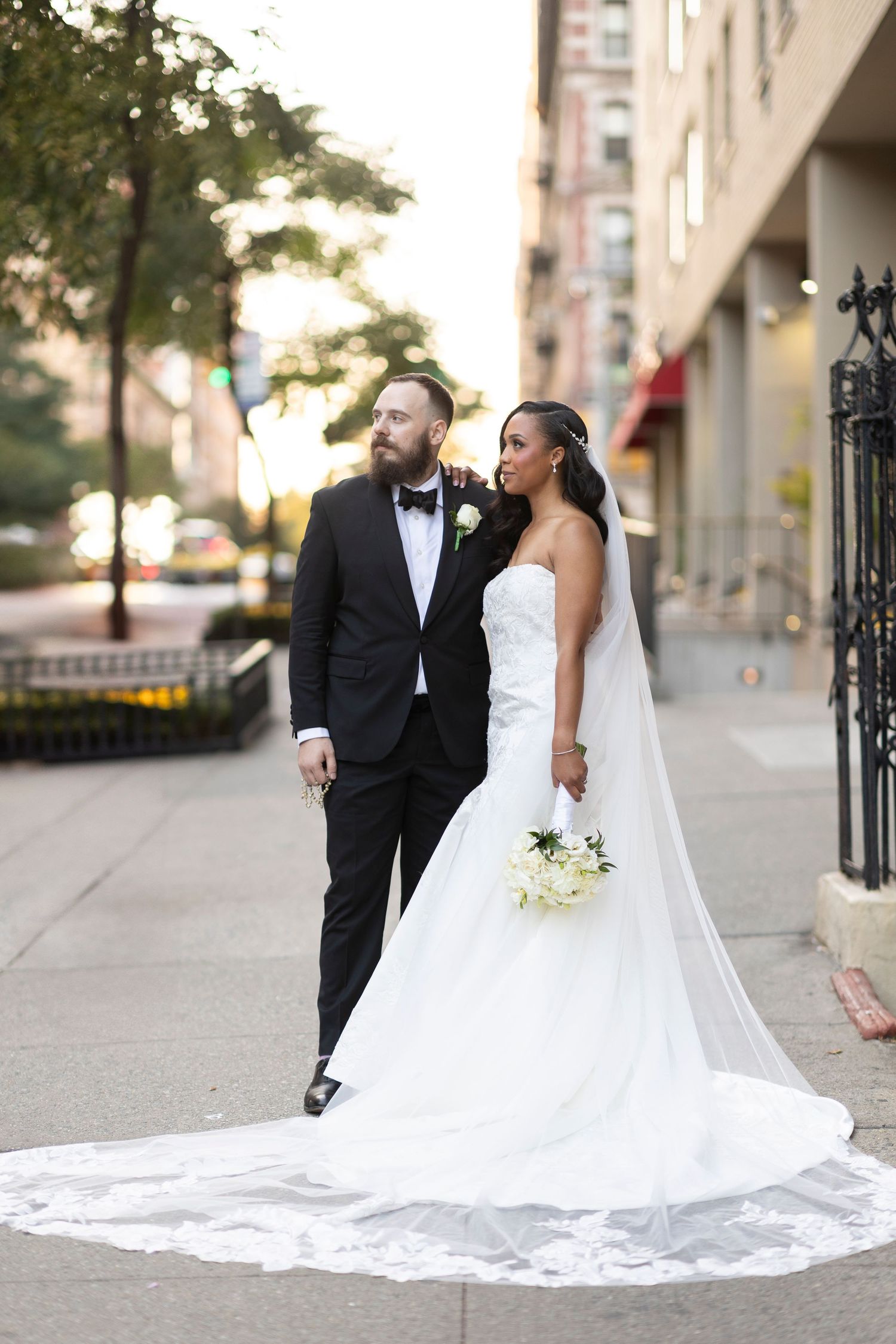Stylish wedding portrait against city backdrop with long white train and classic black tuxedo.