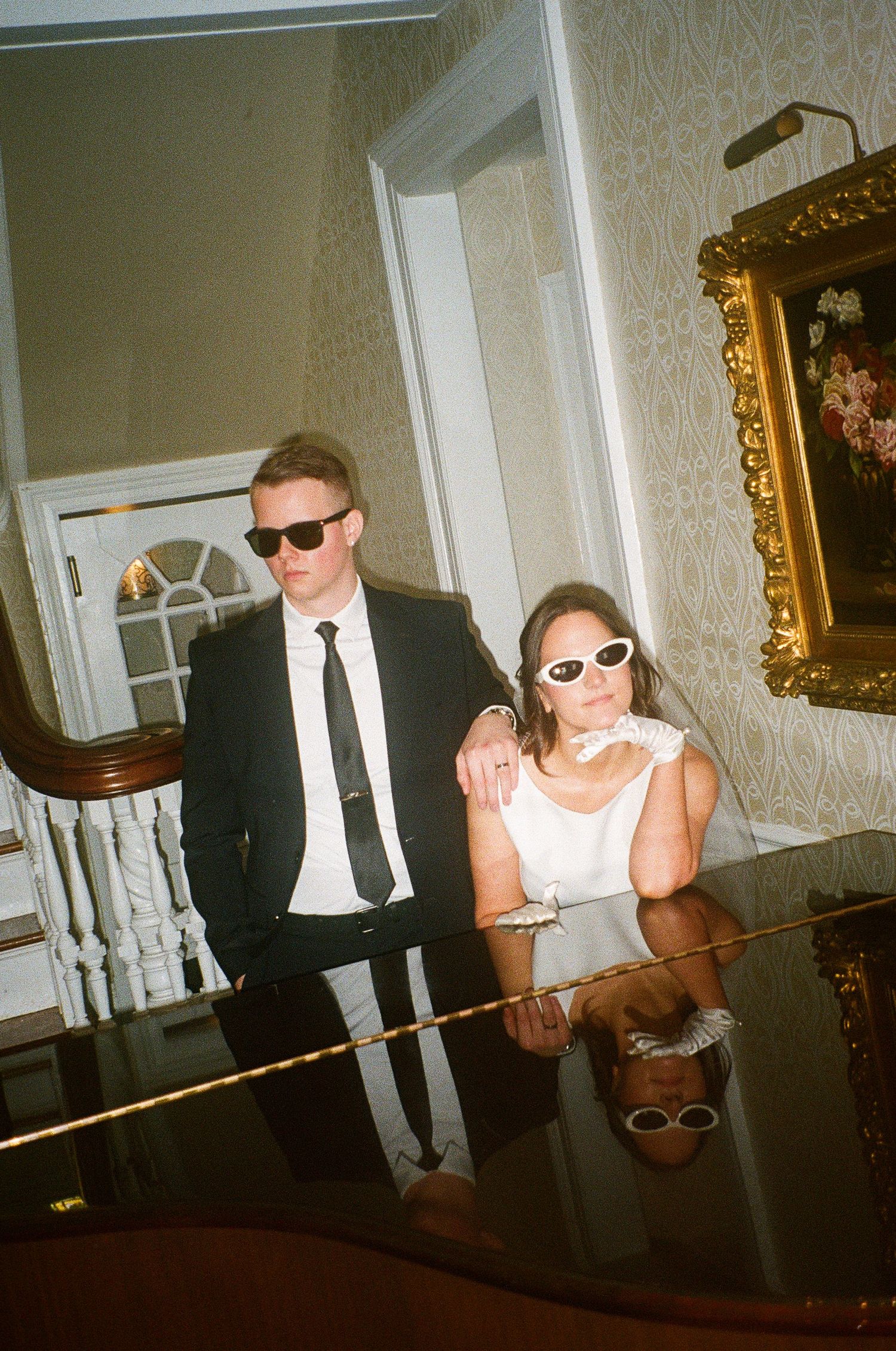 Couple in formal attire wearing sunglasses seated at piano in elegant room.