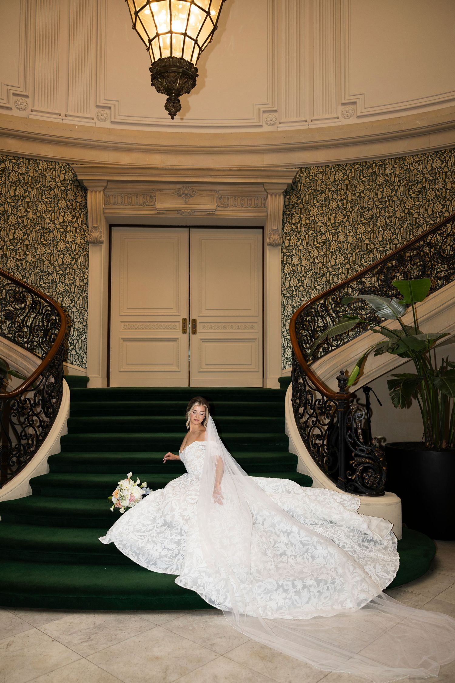 Bride's lace dress spreads elegantly across marble floor near grand staircase.