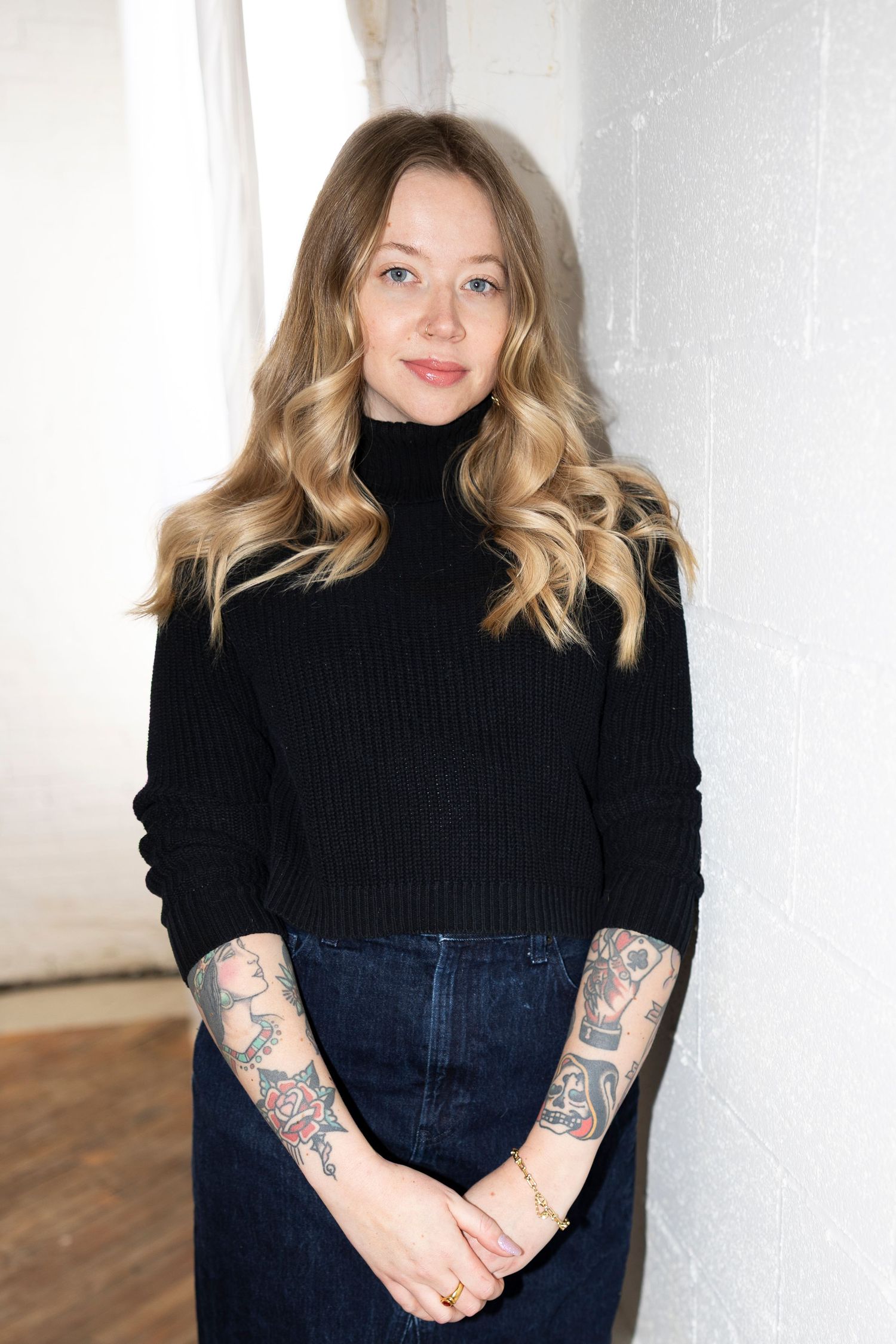 Person wearing black turtleneck sweater and dark jeans shows colorful arm tattoos while posing against white wall.