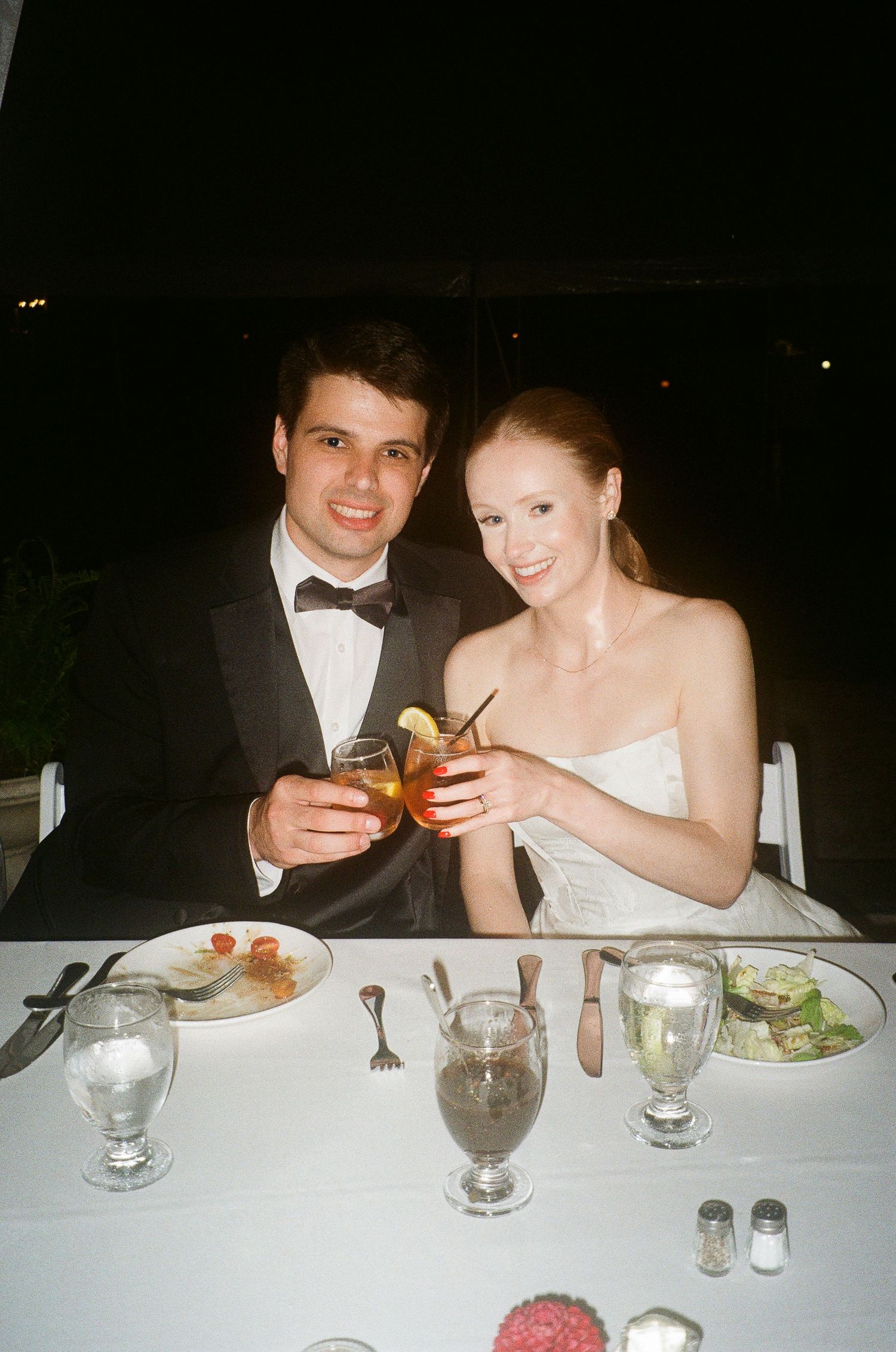 Bride and groom toasting with cocktails at wedding reception dinner table, smiling couple in formal attire.