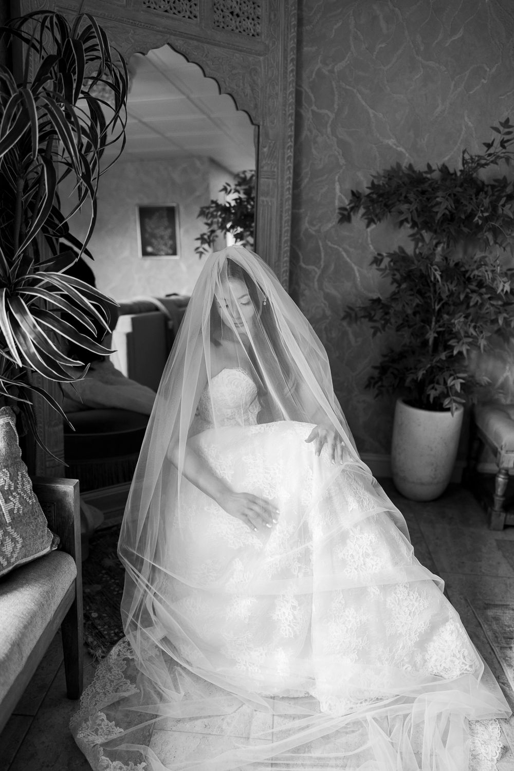 Black and white photo of a bride in a lace gown and flowing veil seated indoors surrounded by plants.