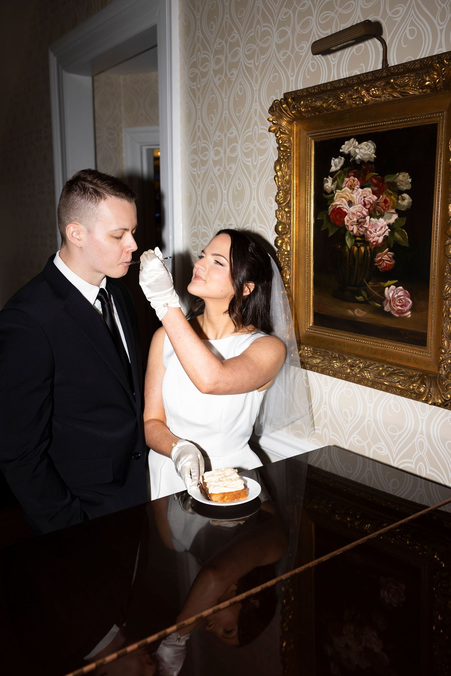 A couple shares cake at their wedding reception in front of an ornate floral painting.