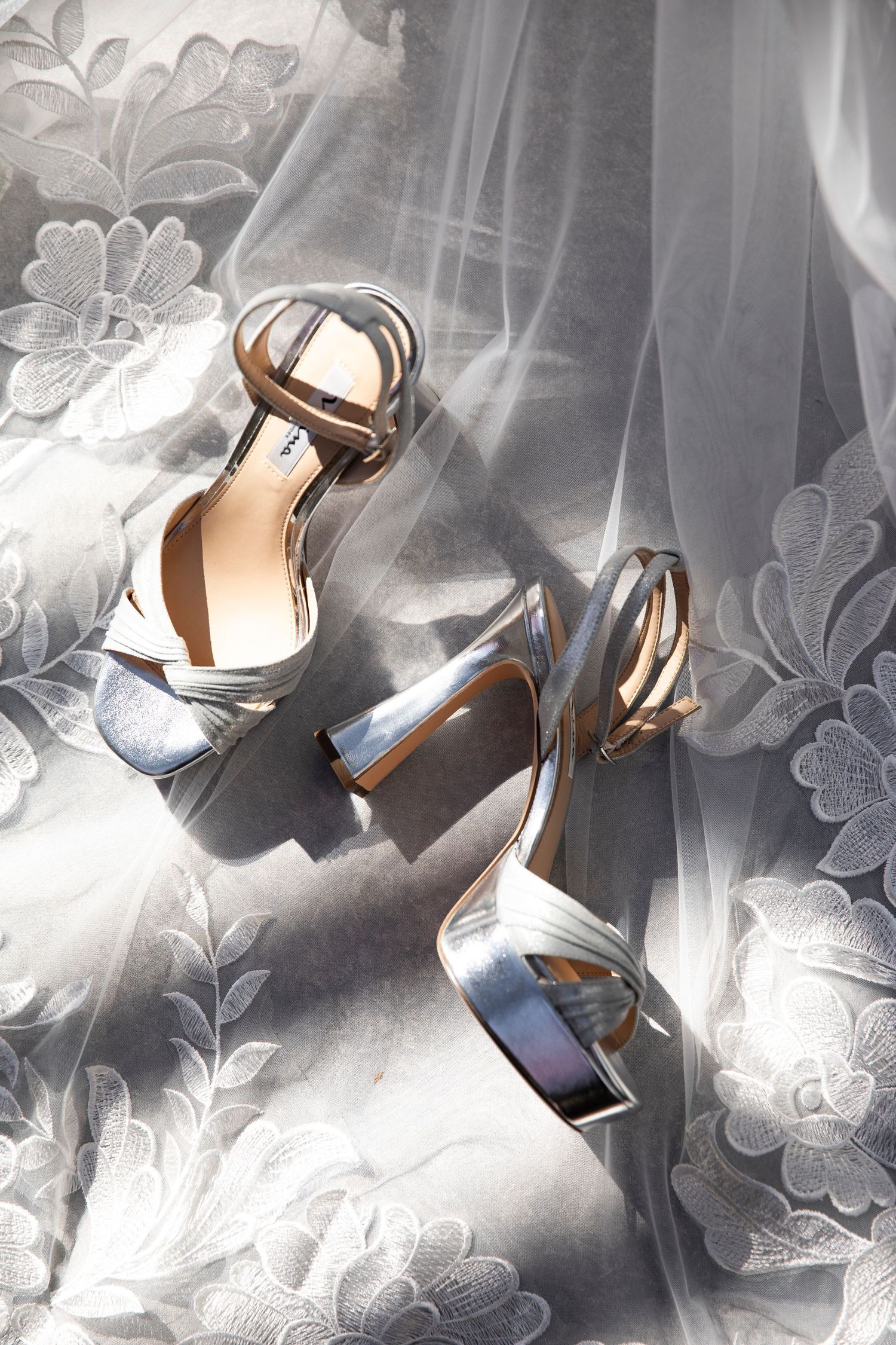 Metallic silver wedding heels with ankle straps photographed on lace fabric.