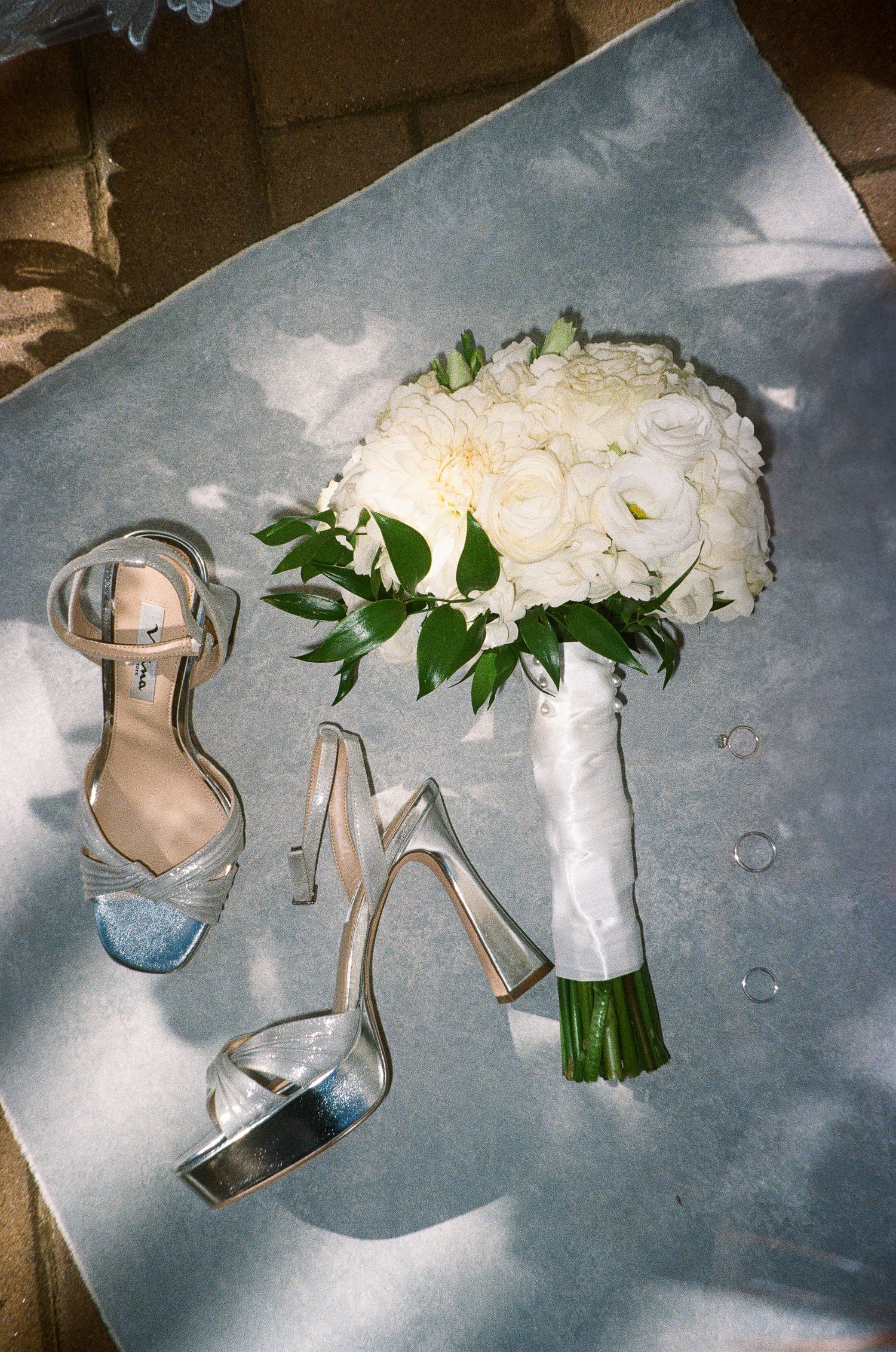 White bridal bouquet with roses and greenery next to silver wedding shoes on reflective surface.