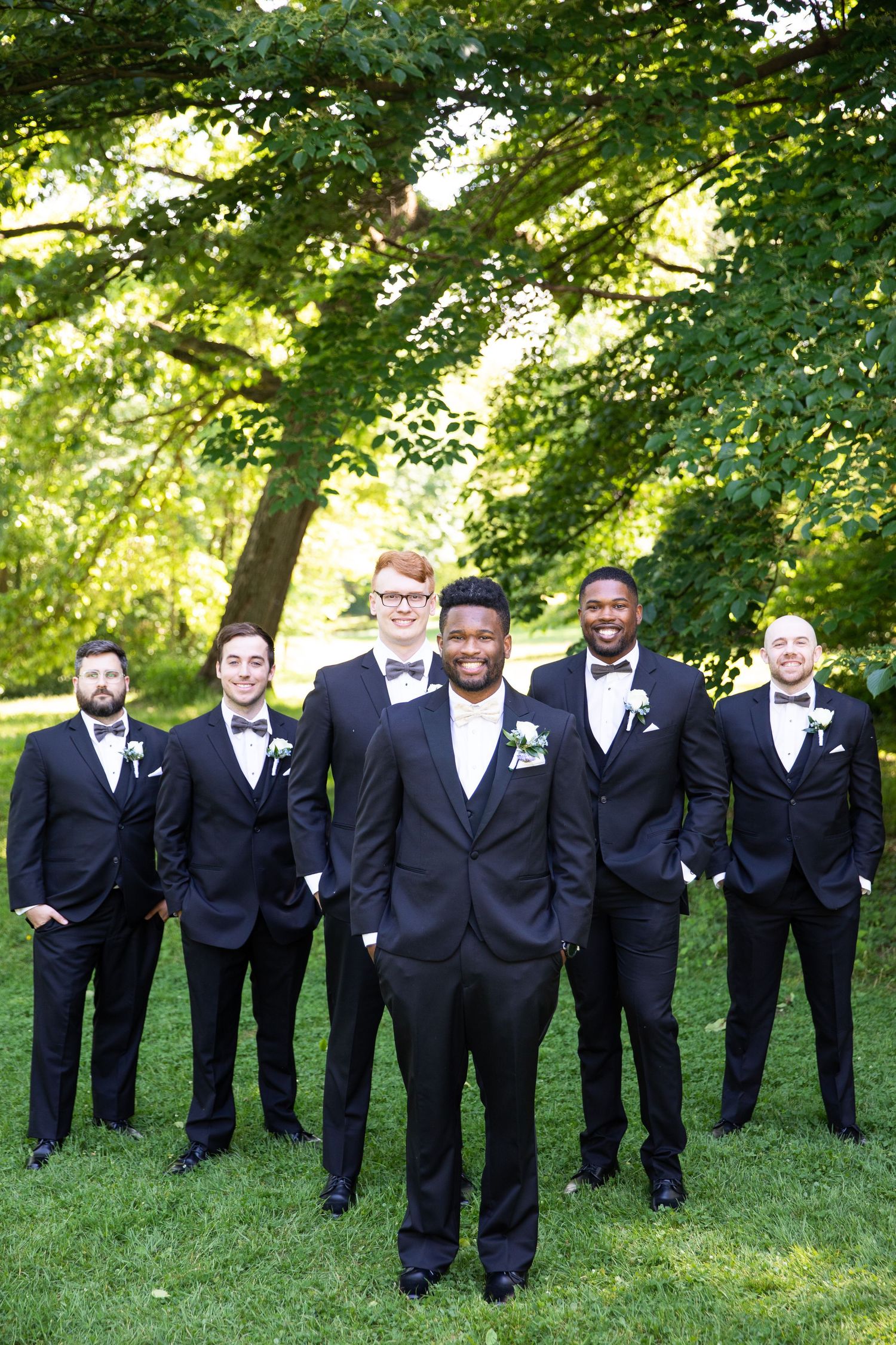 Groomsmen in black tuxedos line up for formal photo on green lawn.