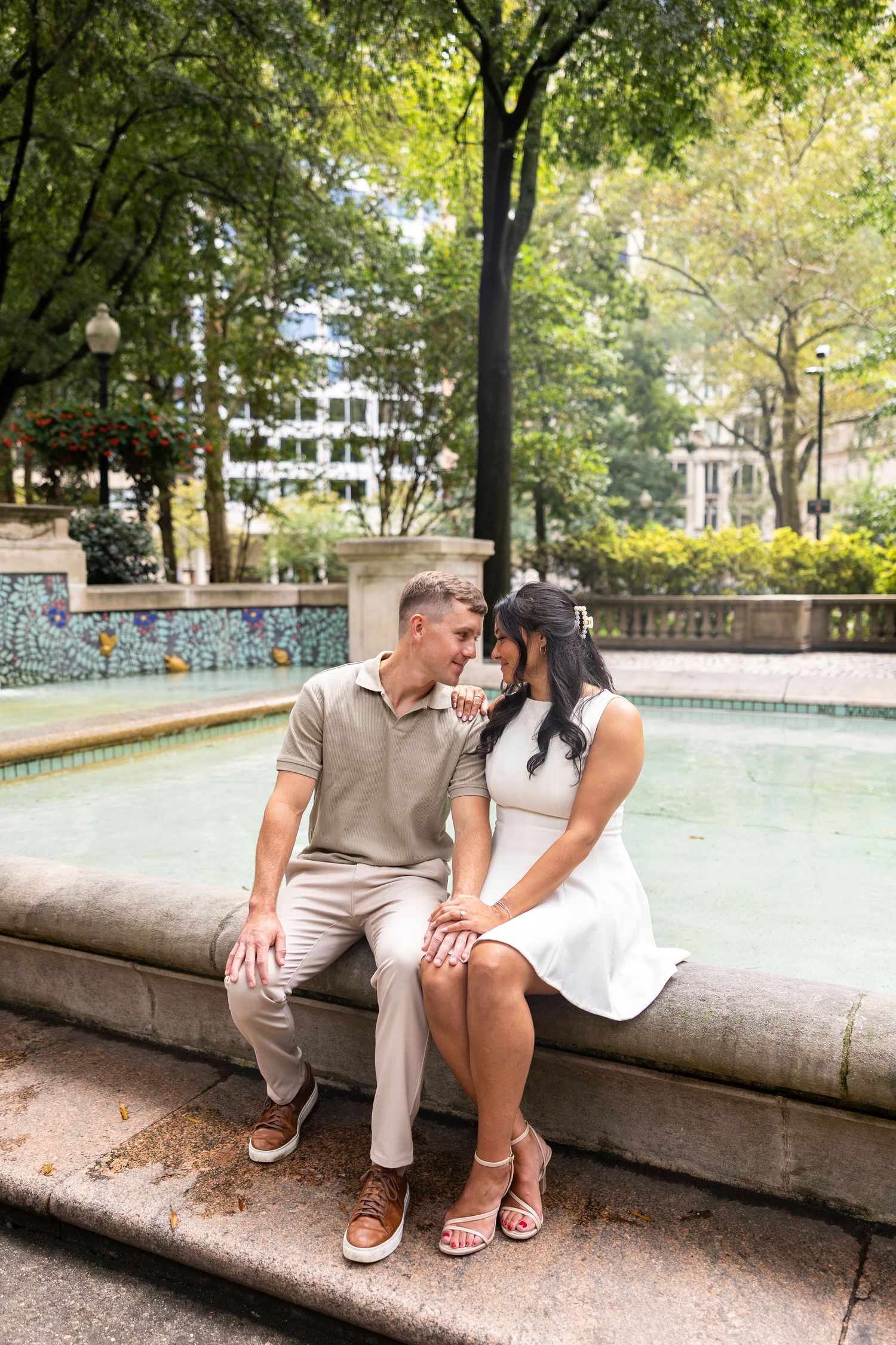 Couple shares intimate moments while sitting on a stone wall near a fountain in a park.