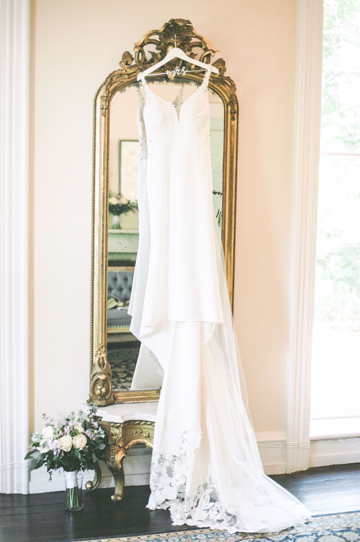 Wedding dress display in a bright room with ornate gold mirrors and natural lighting.