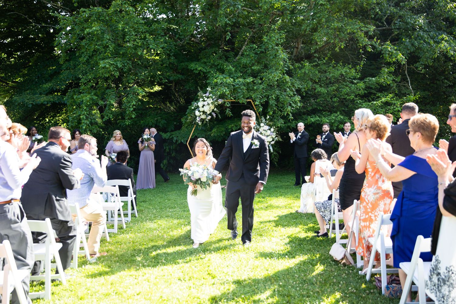 Panoramic view of outdoor wedding ceremony with guests seated on white chairs on grassy hill.