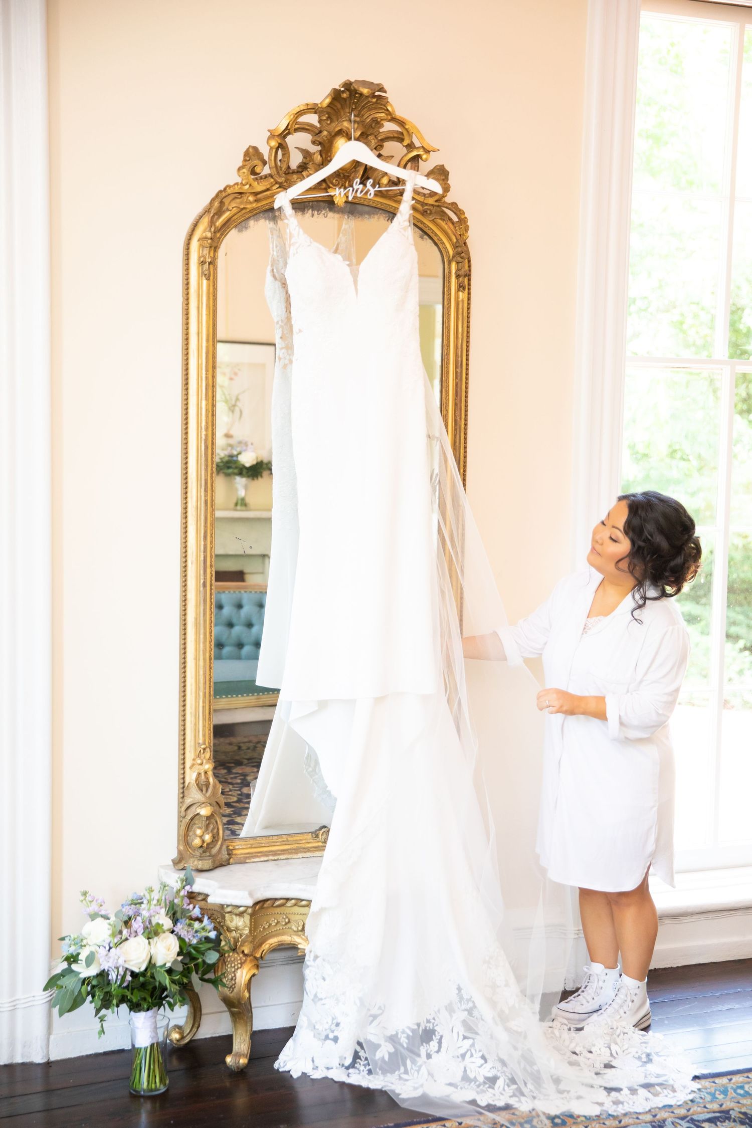A bright room with ornate gold mirrors where a wedding dress hangs near large windows.