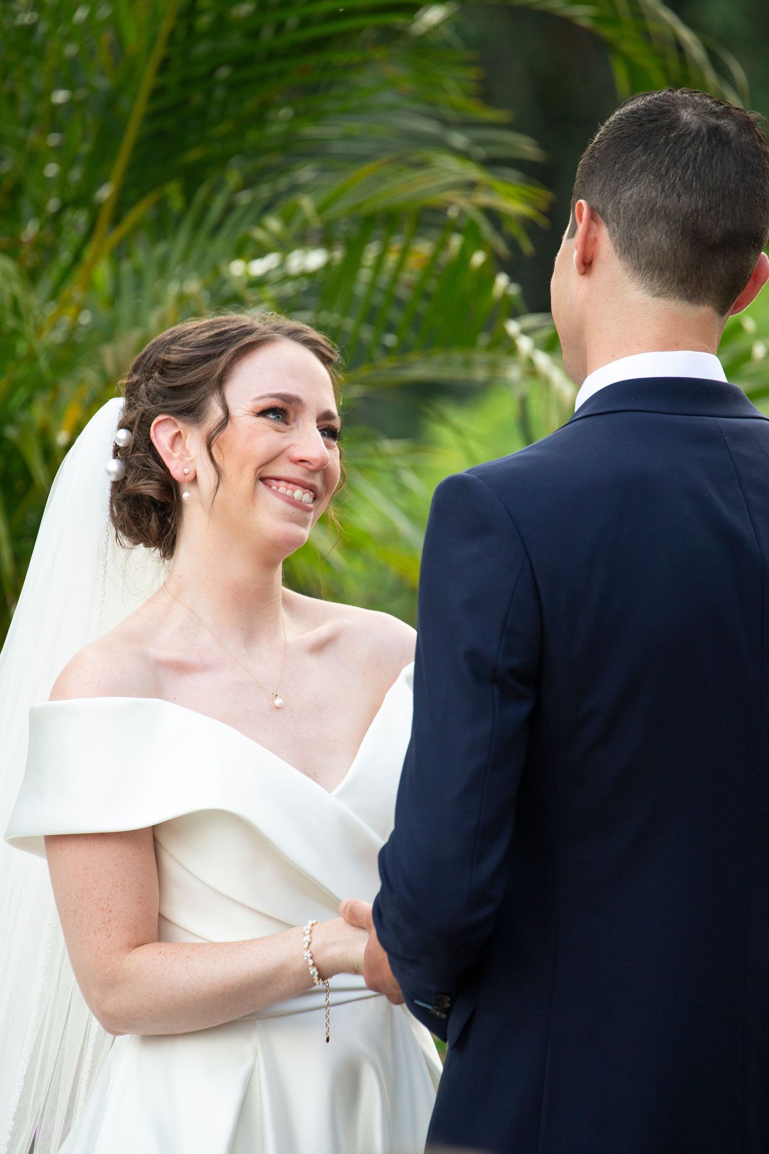 Bride in an elegant off-shoulder wedding dress smiles during outdoor ceremony with tropical foliage.