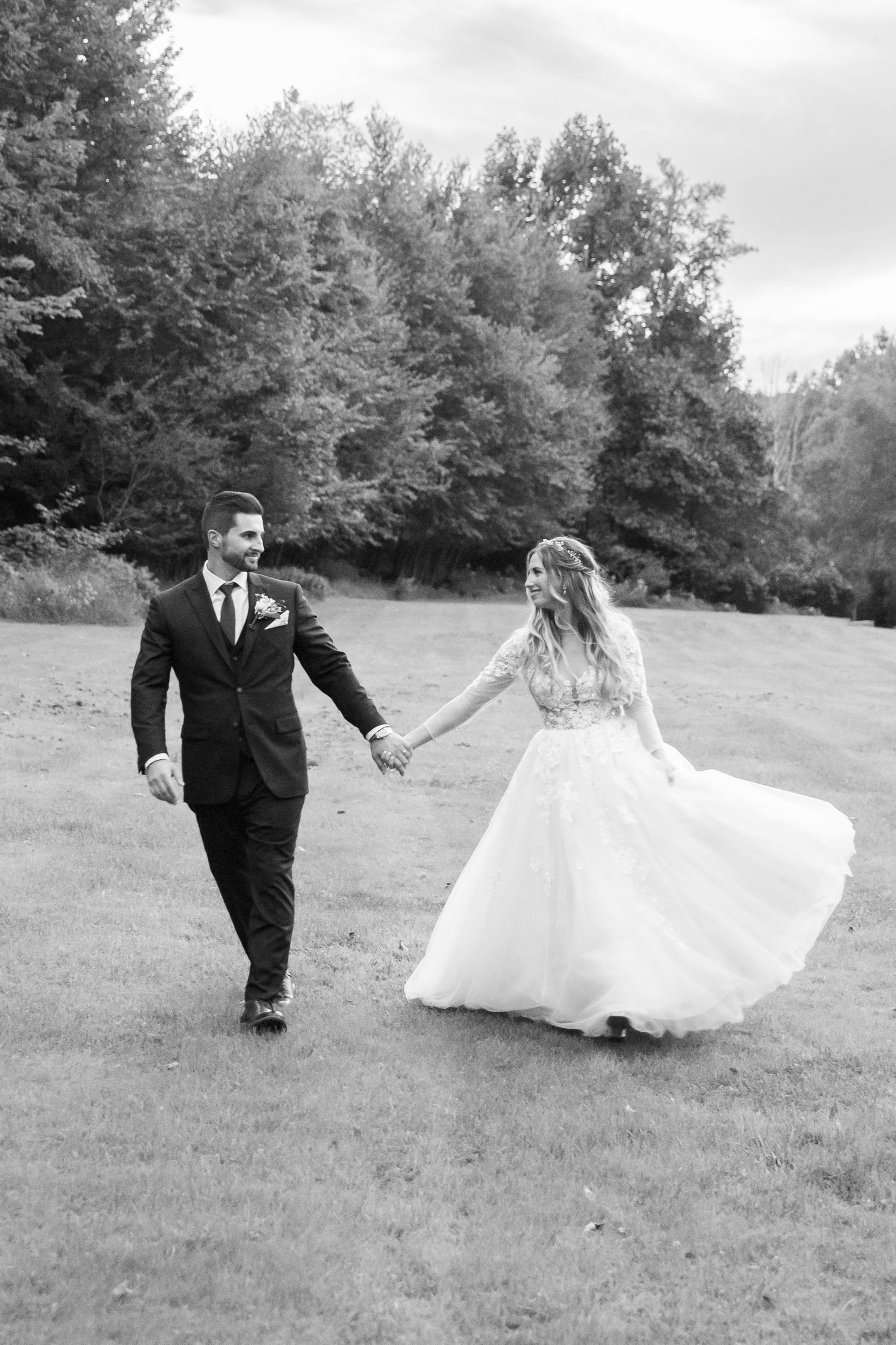 A romantic black and white wedding photo of a couple walking hand in hand across a grassy field.