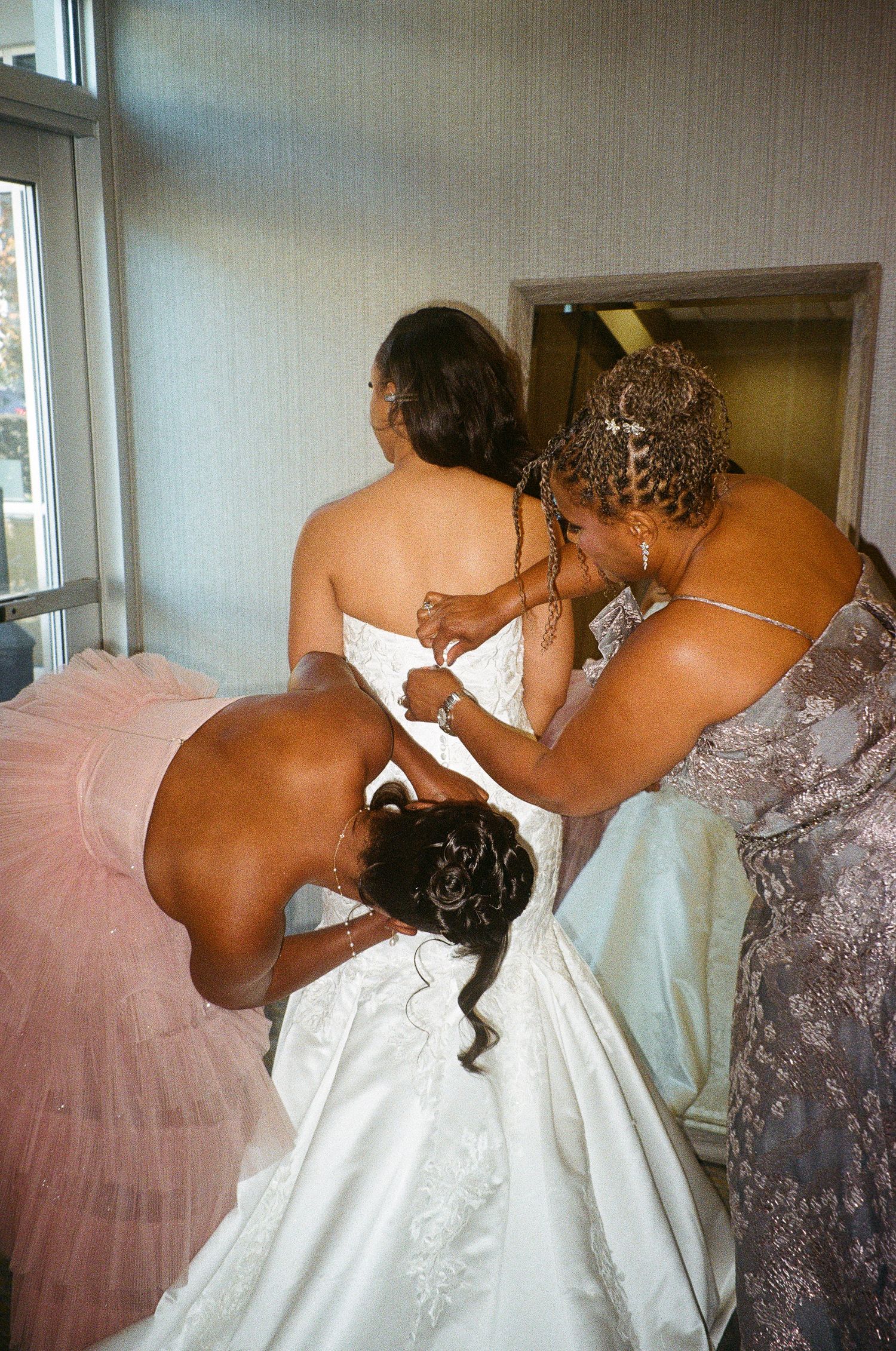 Helpers assisting with fastening the back of an ornate white wedding gown.