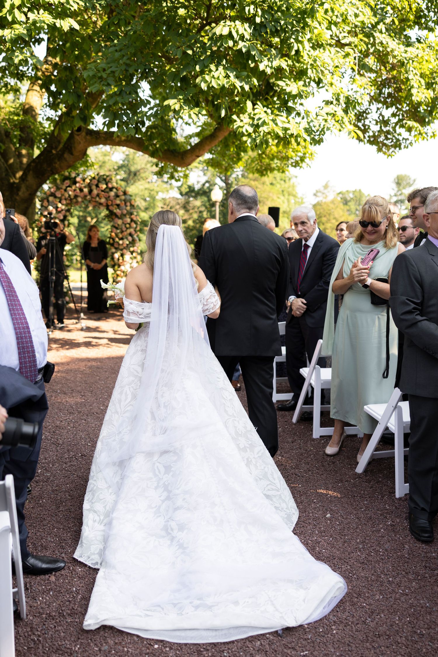 Wedding guests seated in white chairs on a rustic outdoor path lined with trees.