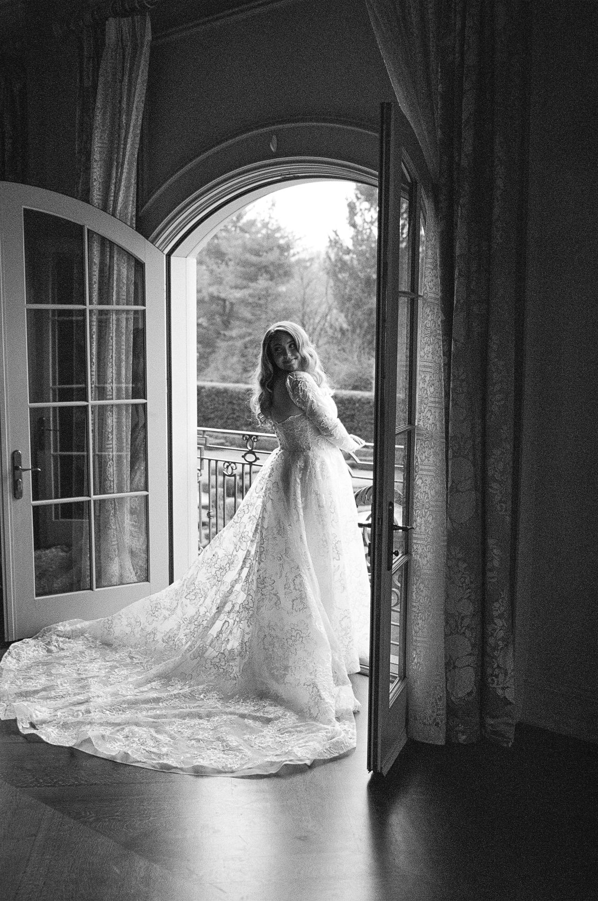 Bride in lace wedding gown with long train stands in arched French doors, black and white photo.