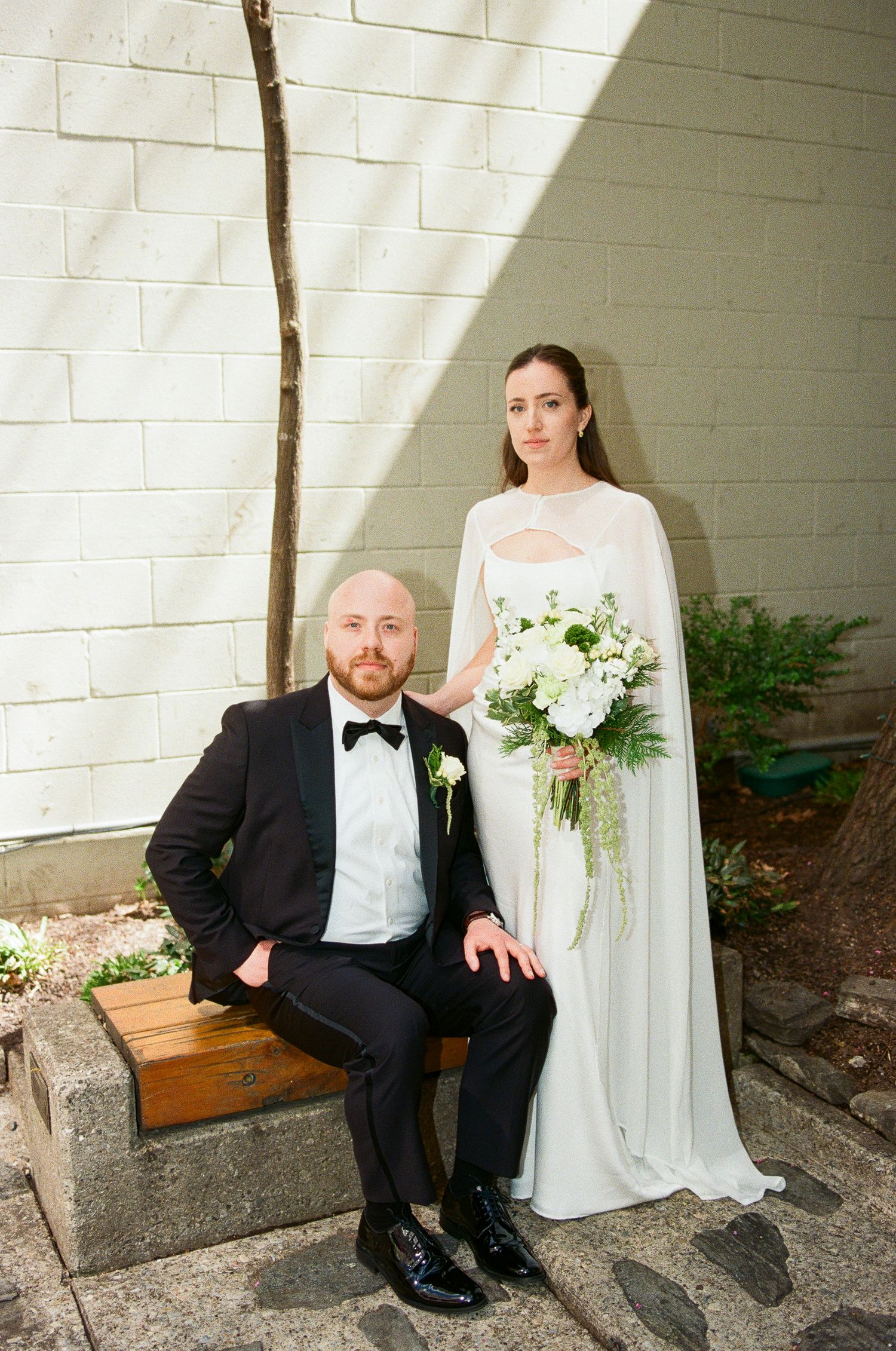 Elegant bride in white cape gown holding green bouquet stands beside groom in black tuxedo seated on bench outdoors.