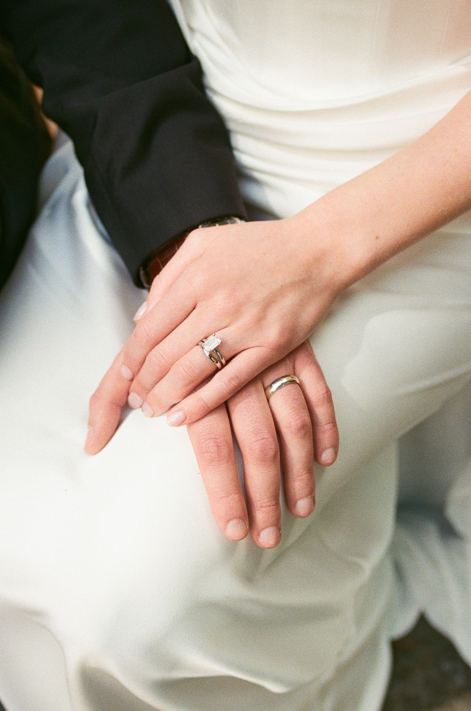Close-up of bride and groom hands showing wedding rings on white dress.