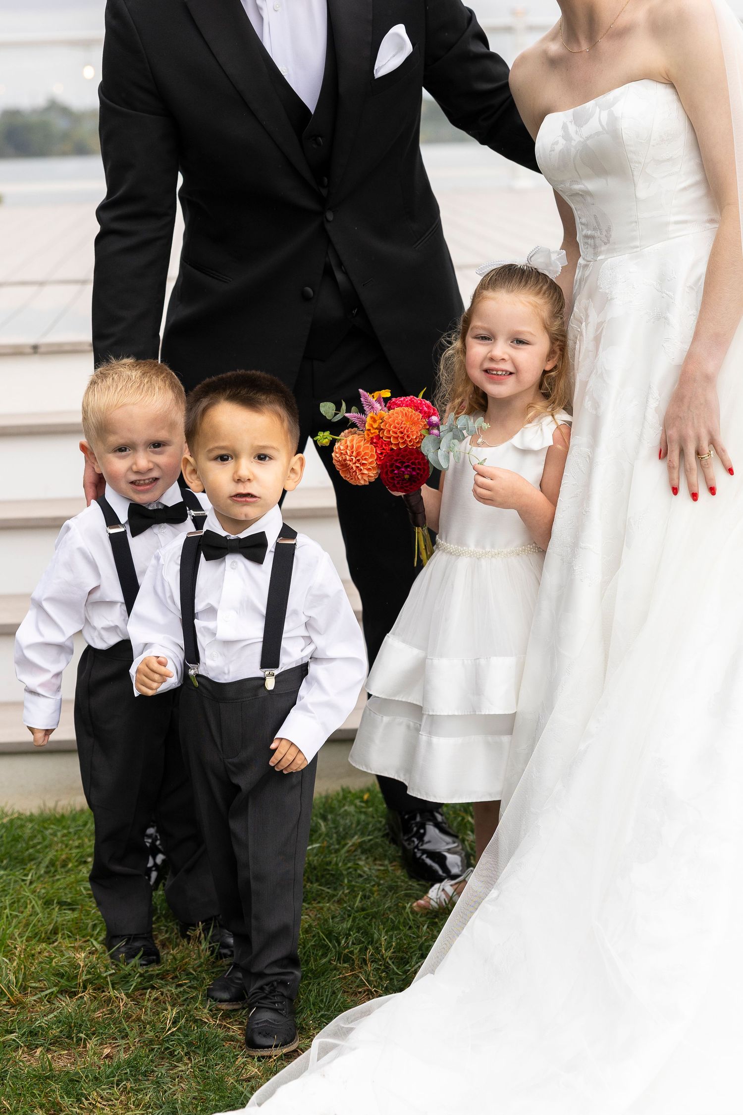 Two ring bearer boys and a flower girl in formal attire stand with bride and groom at an outdoor wedding.