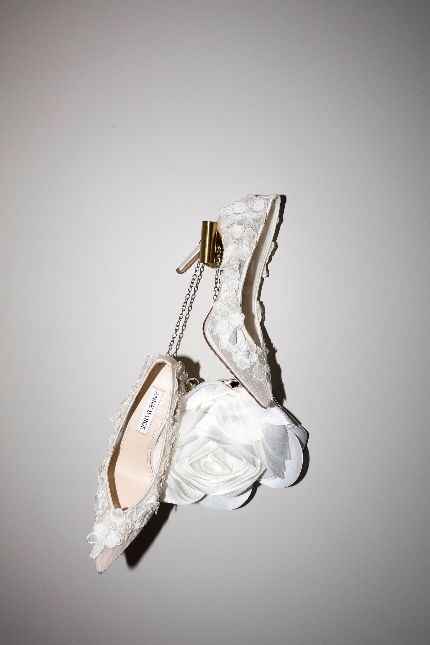 White lace bridal heels with floral embellishments and gold ankle chain, adorned with a satin rose, suspended mid-air.