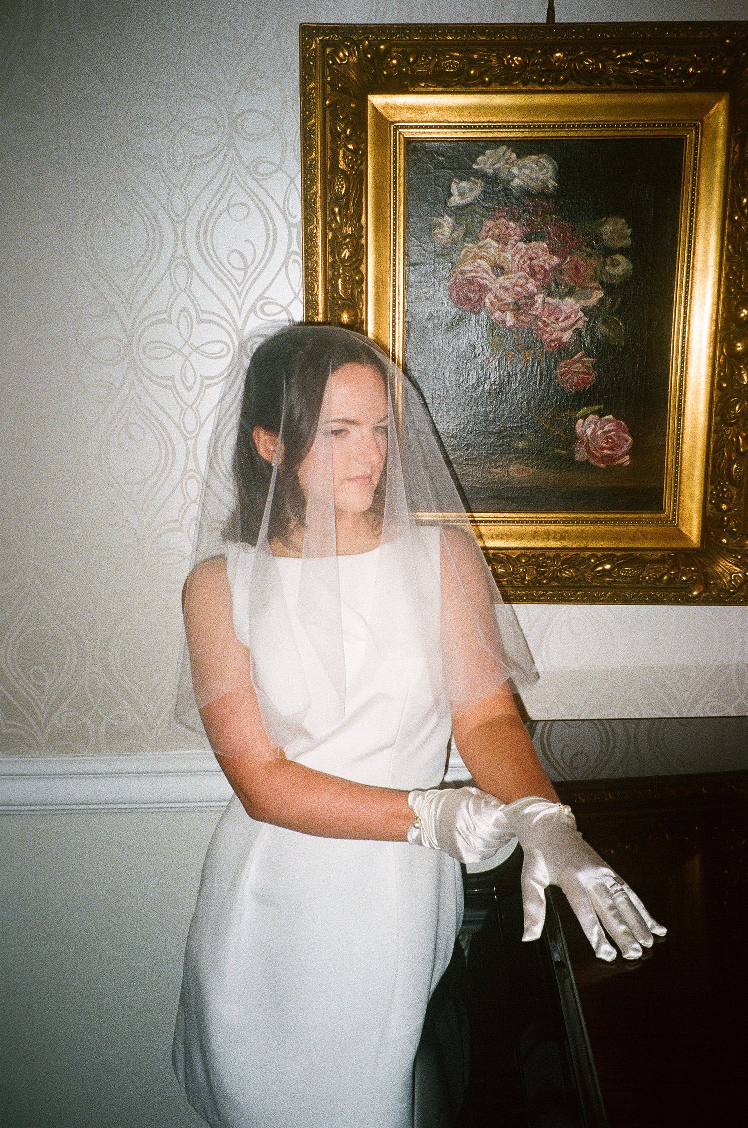 A bride in a white dress and veil poses against a wall with ornate gold-framed artwork.