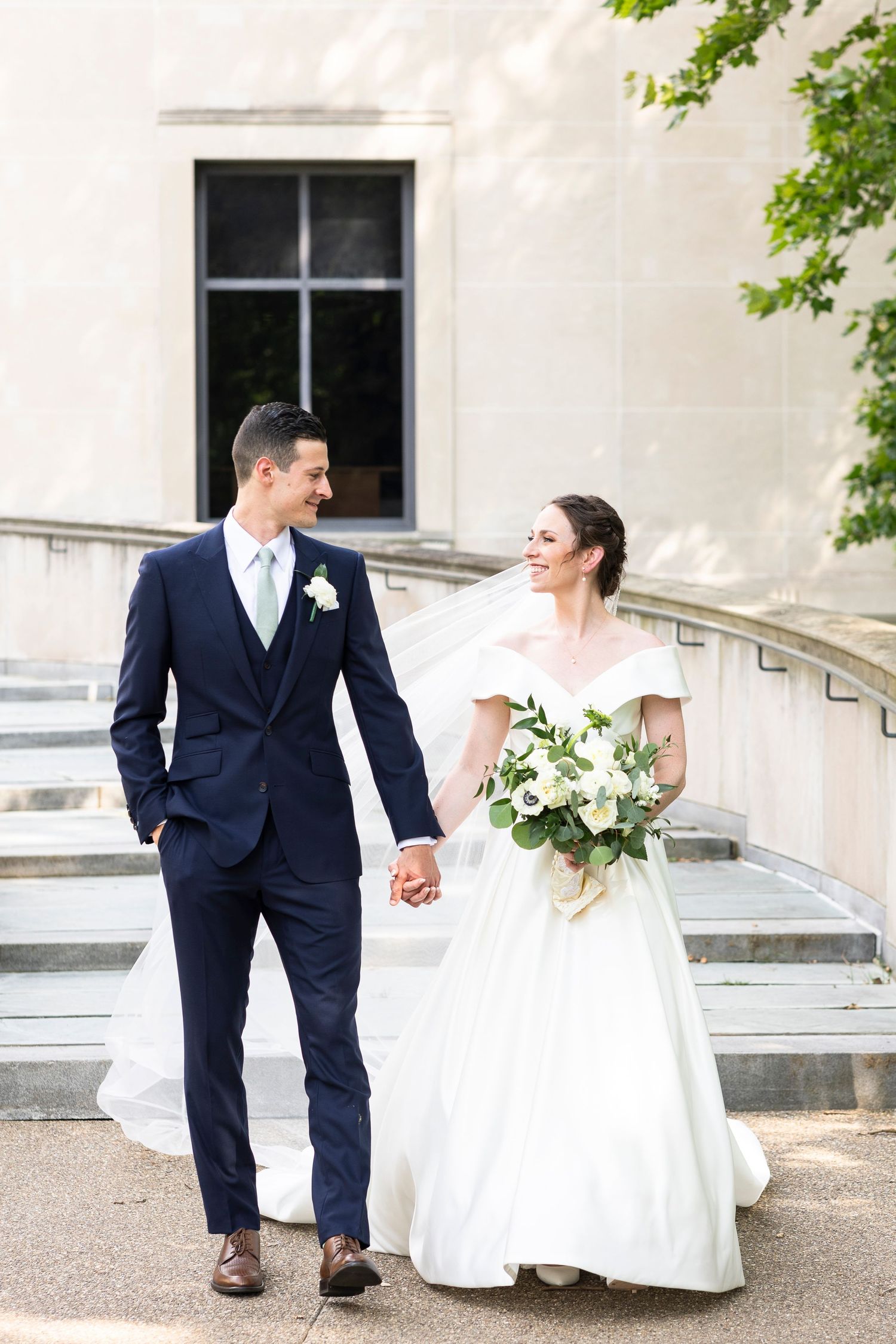 Newlyweds walking down stone steps with white bouquet and navy suit against classic architecture.
