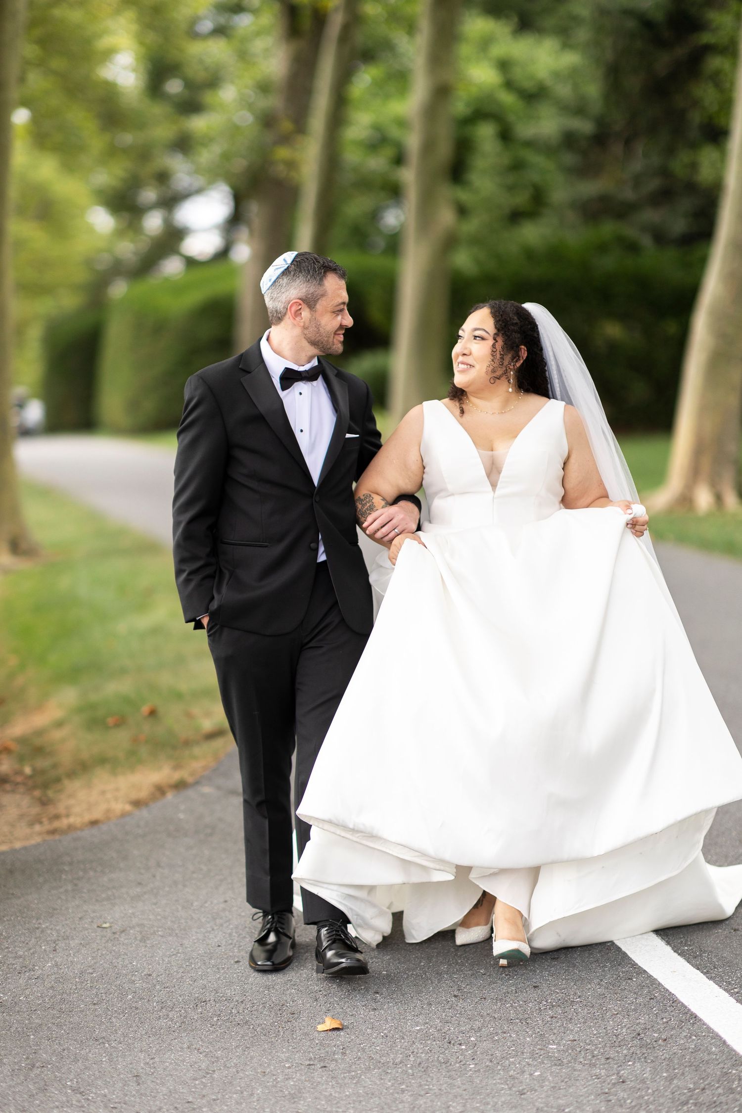 A joyful wedding couple walking together on a tree-lined path while the bride holds her flowing white dress.