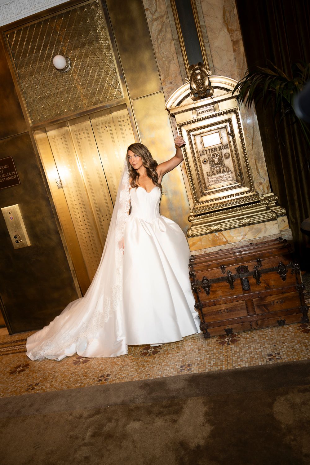 Bride in elegant white ball gown with veil poses near gold elevator and ornate mailbox in luxurious hotel lobby.