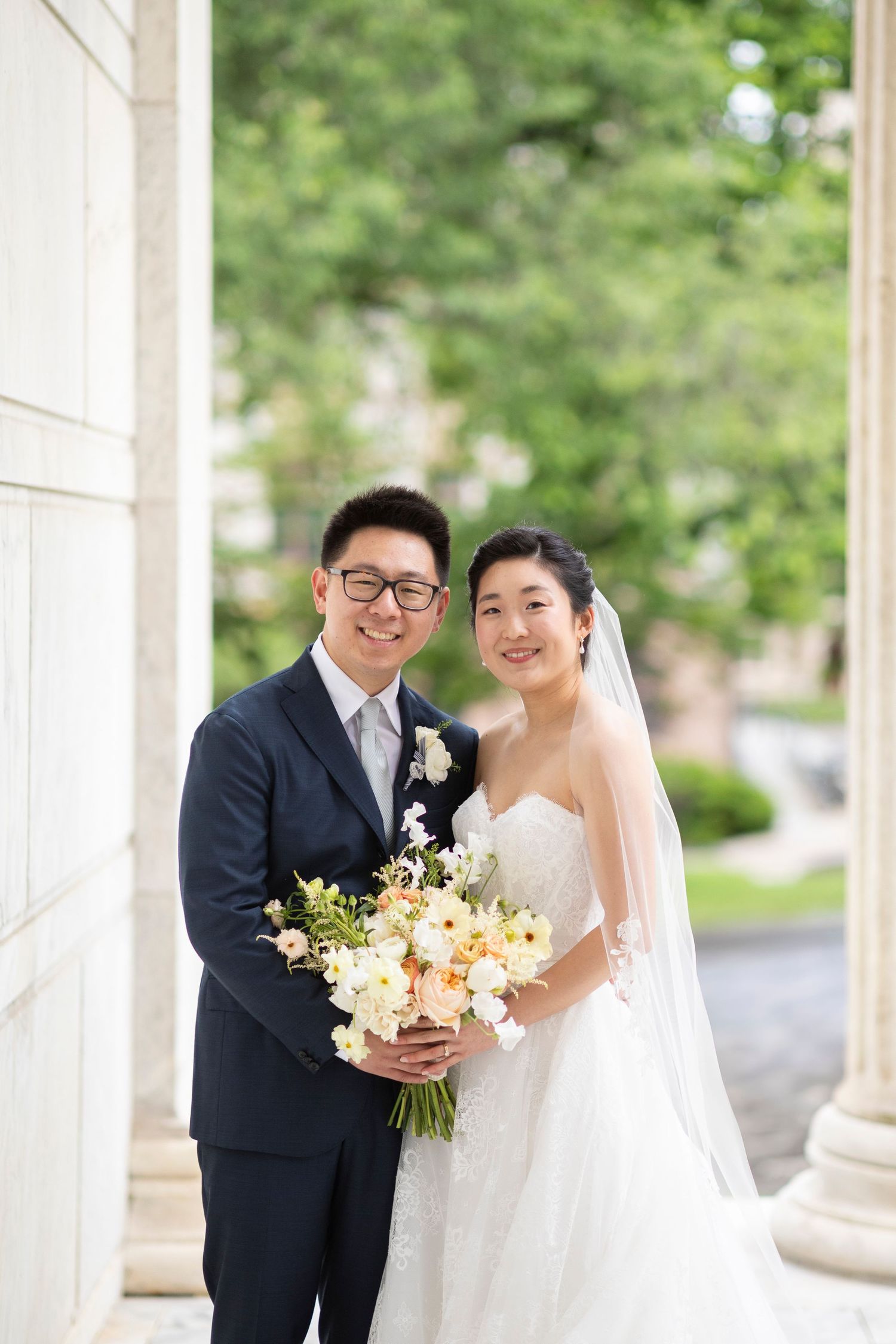Smiling bride and groom pose with white floral bouquet between elegant columns outdoors on their wedding day.