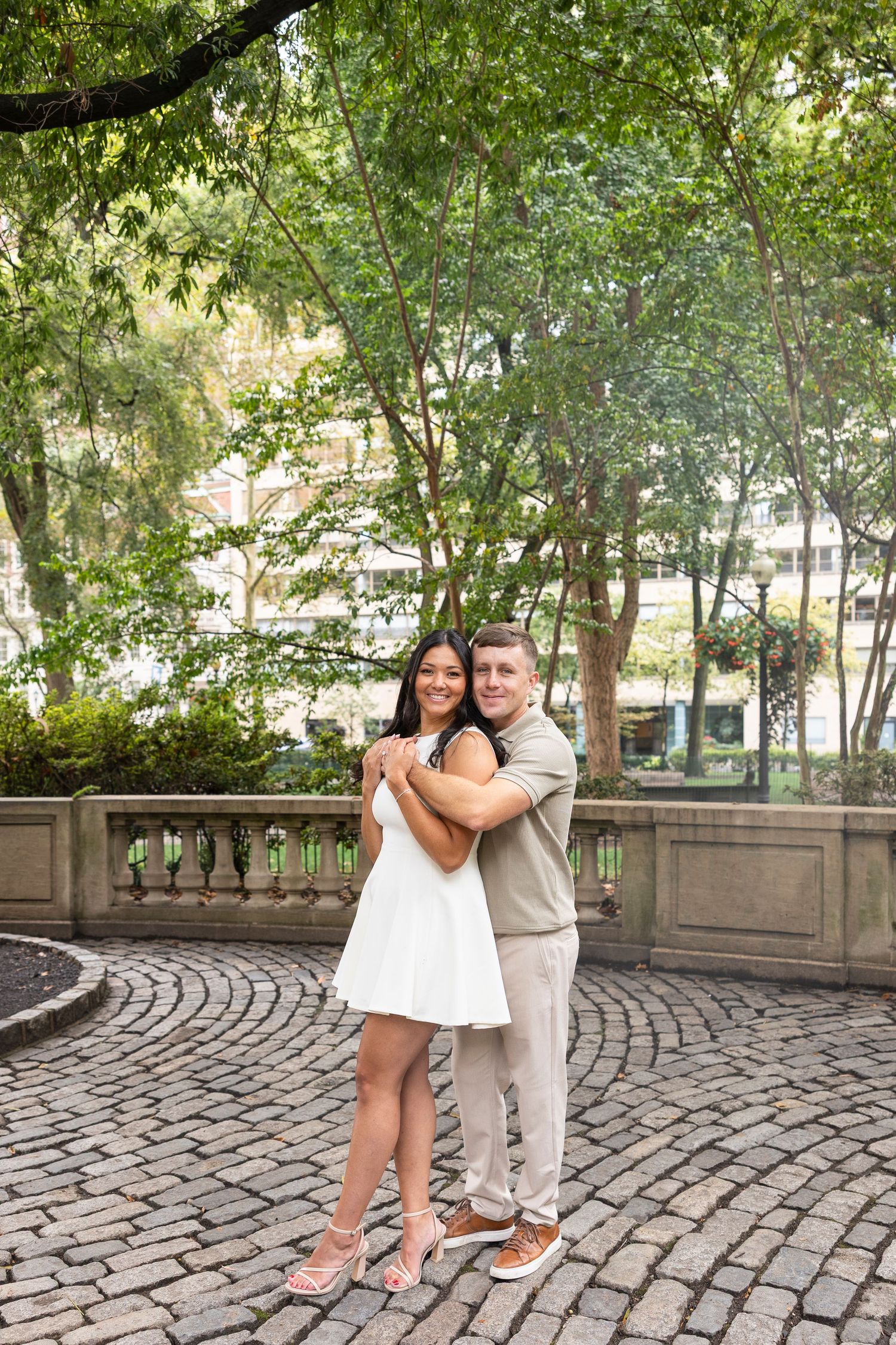 A couple poses on cobblestone pavement surrounded by lush green trees.