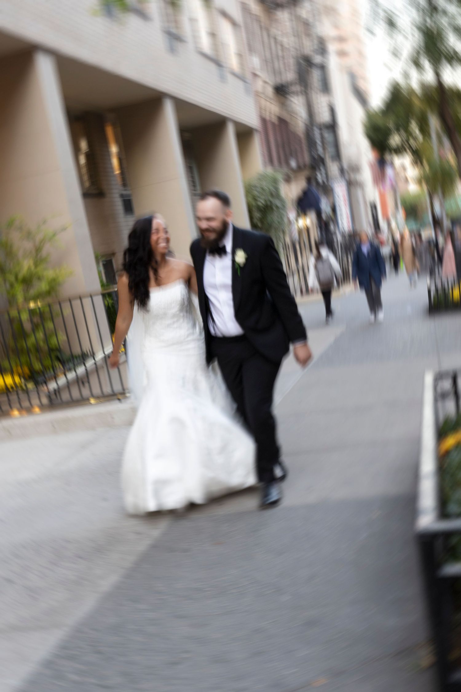 Motion-blurred candid wedding photo of couple walking down city street.
