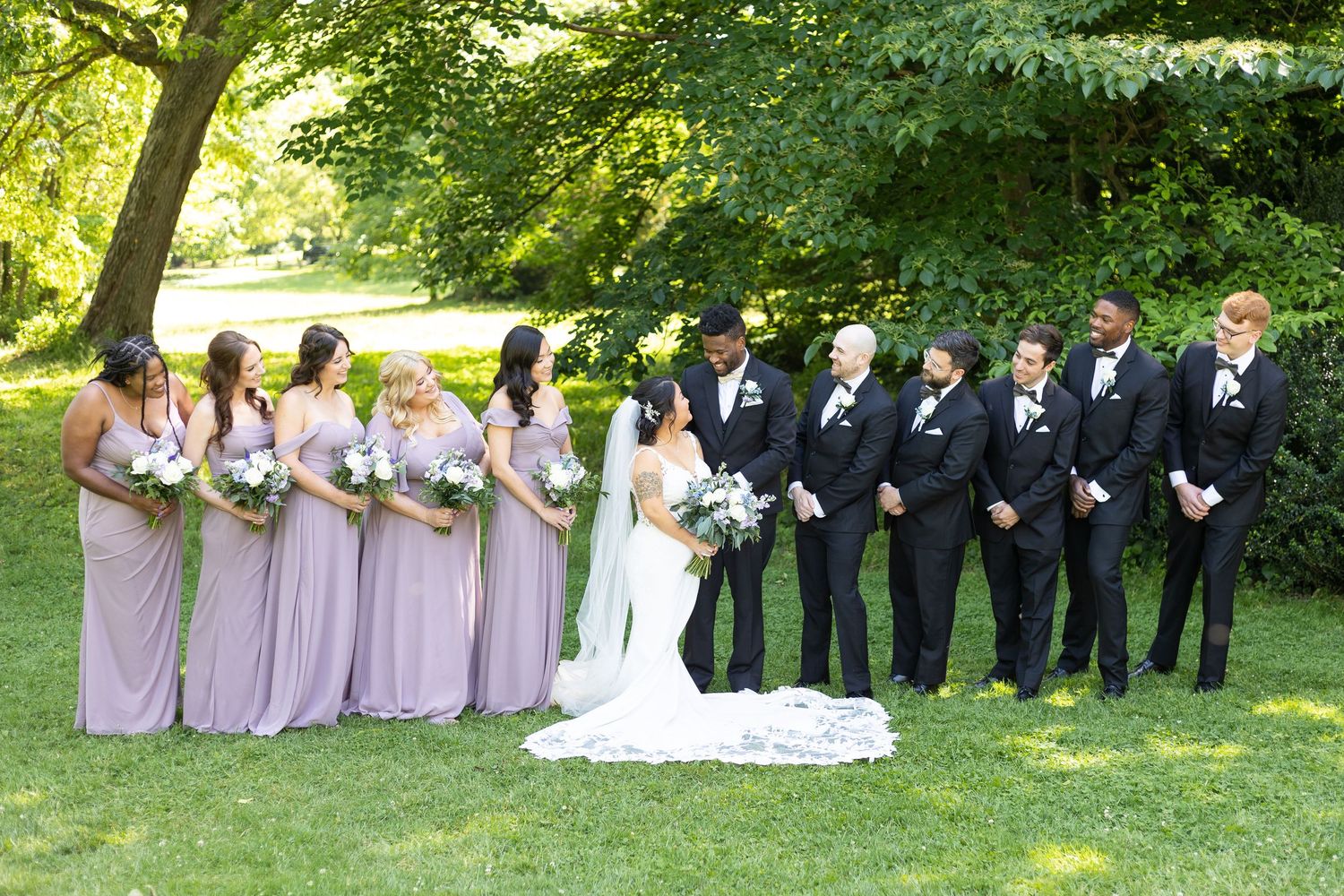 Wedding party in formal attire poses together on lush green lawn under tree canopy.