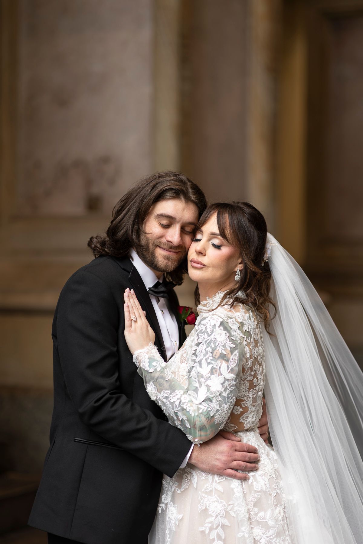 Bride and groom embracing tenderly in elegant wedding attire, lace dress with veil, black tuxedo, romantic indoor setting.