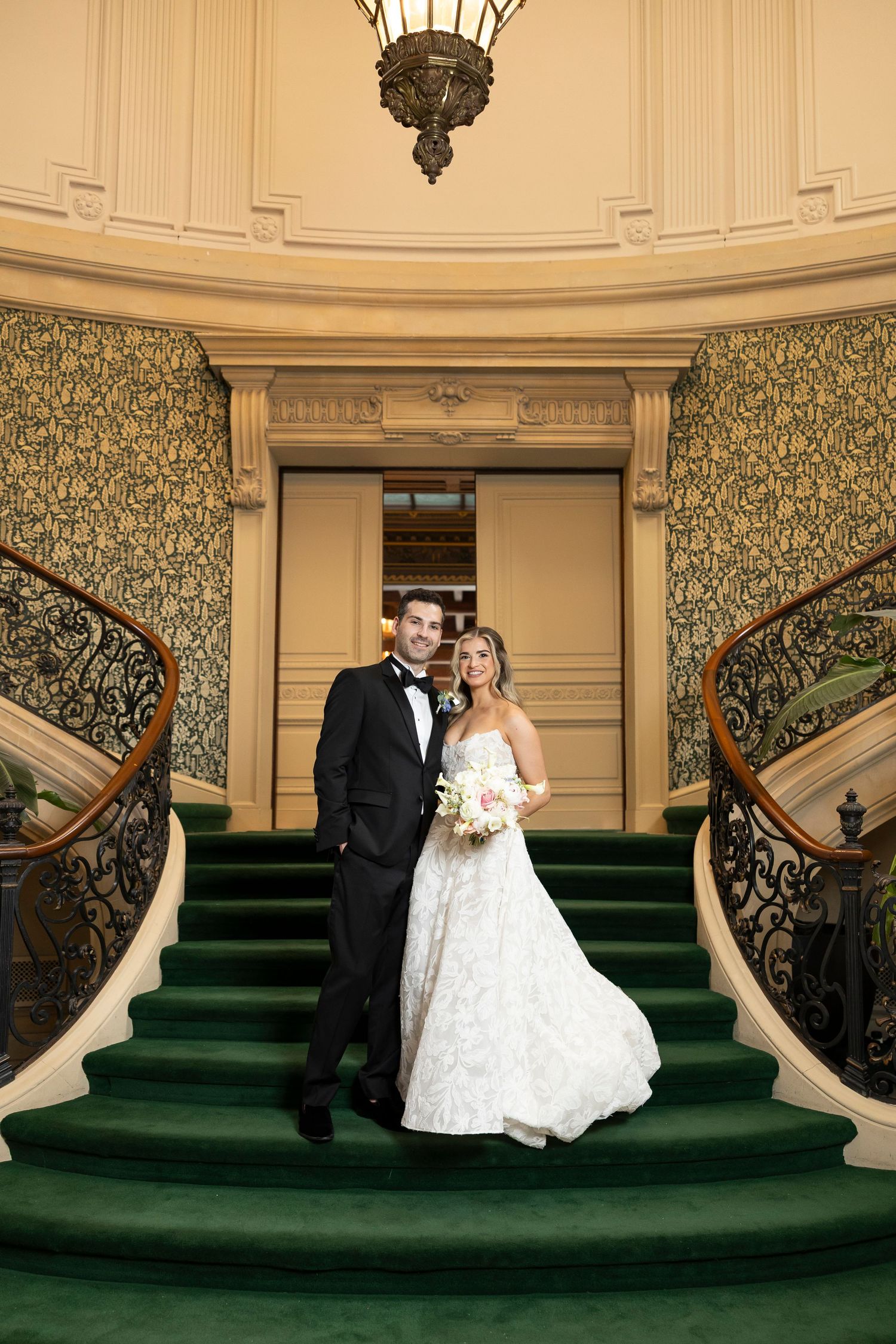 Wedding couple poses on grand staircase with green carpet and ornate railings.