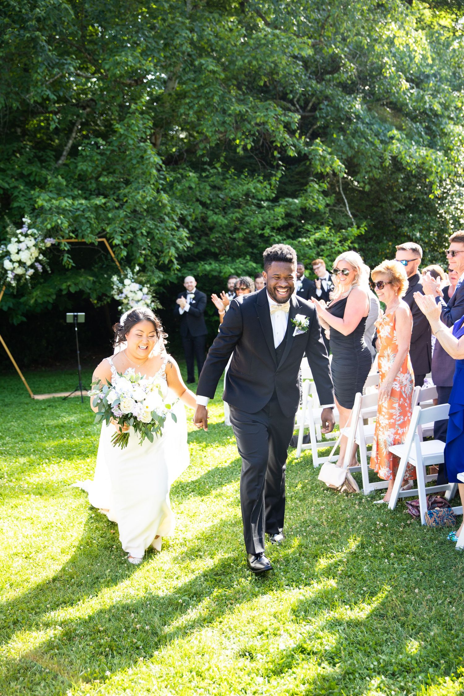 Wedding ceremony guests seated on white chairs in a sunny outdoor garden setting.